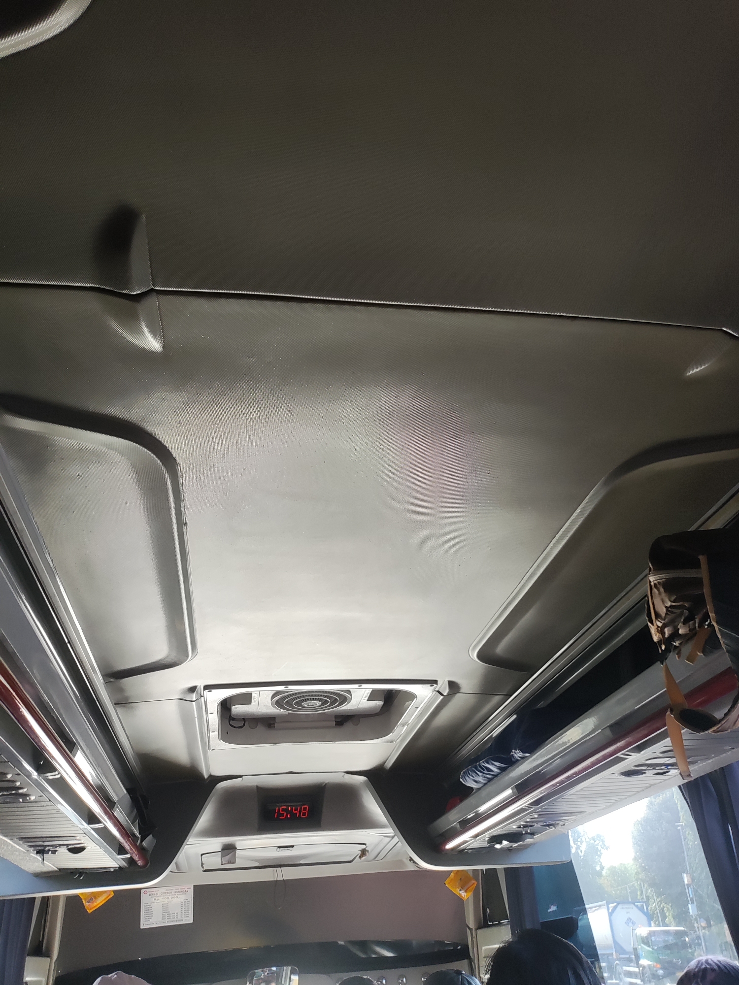The image captures the interior of a bus, focusing on the ceiling and overhead compartments. A digital clock displays "15:48". Luggage is stored in the overhead bins. Outside the bus, through the windows, trees and a truck are visible, suggesting the bus is in motion. The setting is likely Purwakarta, Indonesia, given the context provided.