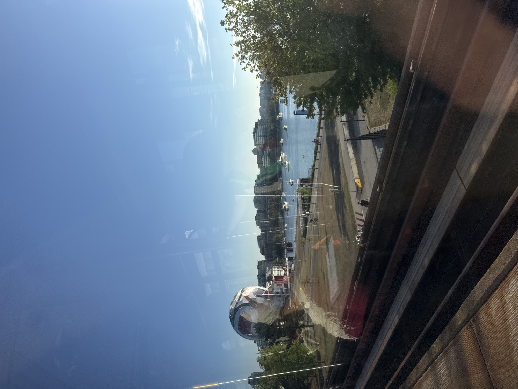 This image captures a view of the False Creek area in Vancouver, Canada, as seen through a window. In the foreground, a set of parallel metal tracks, possibly from a SkyTrain, dominate the lower portion of the frame, with a textured surface beneath them. Beyond the tracks, a grassy area leads to a waterfront promenade.
Dominating the left side of the mid-ground is a large, geodesic dome structure resembling a soccer ball, known as the TELUS World of Science. Several trees, some with lush green foliage, are scattered throughout the park area. The calm waters of False Creek are visible in the background, dotted with numerous white boats and yachts. A modern city skyline with several multi-story buildings forms the backdrop across the water.
The sky is a clear, vibrant blue with a few wispy white clouds. The light suggests it is either early morning or late afternoon, with the sun casting a warm glow. Reflections are visible on the window, including a blurry red object on the left and lines that suggest the window panes.
In the mid-ground, a few individuals are present. On the promenade, two people are walking, and a cyclist is visible further down. A lone figure is seen walking on the pathway near the water's edge. The scene depicts a tranquil urban environment, blending recreational spaces with city infrastructure and the natural beauty of the waterfront.