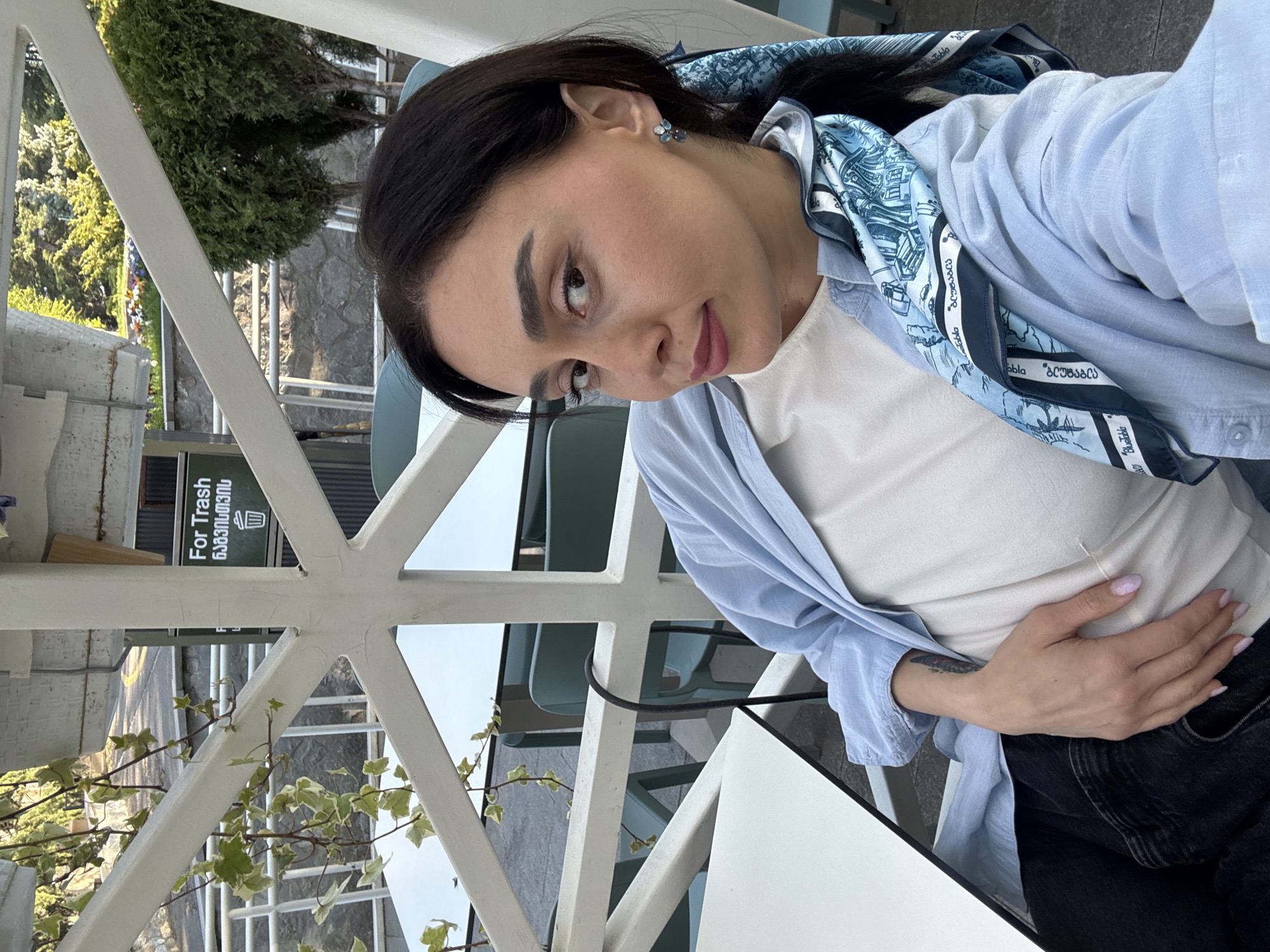 A woman is captured in a selfie-style shot, seated outdoors, likely at a cafe or restaurant in Tbilisi, Georgia. She has dark hair and is wearing a light blue button-up shirt over a white top, paired with dark pants. A blue and white patterned scarf, featuring what appear to be architectural sketches and Georgian text including "ქუთაისი" (Kutaisi) and "ბაგრატ" (Bagrat), is draped around her shoulders. She wears small, blue, flower-shaped earrings. Her left hand rests on her midsection, revealing a light-colored manicure and a small tattoo on her forearm.
The setting includes white tables and part of a white metal lattice structure, possibly a pergola or balcony railing. Light blue chairs are partially visible in the background. Beyond the railing, green trees, ivy, and a stone wall indicate an outdoor, possibly elevated, location with a view of a street or pathway. The time of day appears to be bright daytime.
A black and white sign in the background clearly reads "For Trash" with the Georgian translation "ნაგვისთვის" below it, reinforcing the location context of Georgia. The woman's expression is calm and neutral, looking towards the camera.