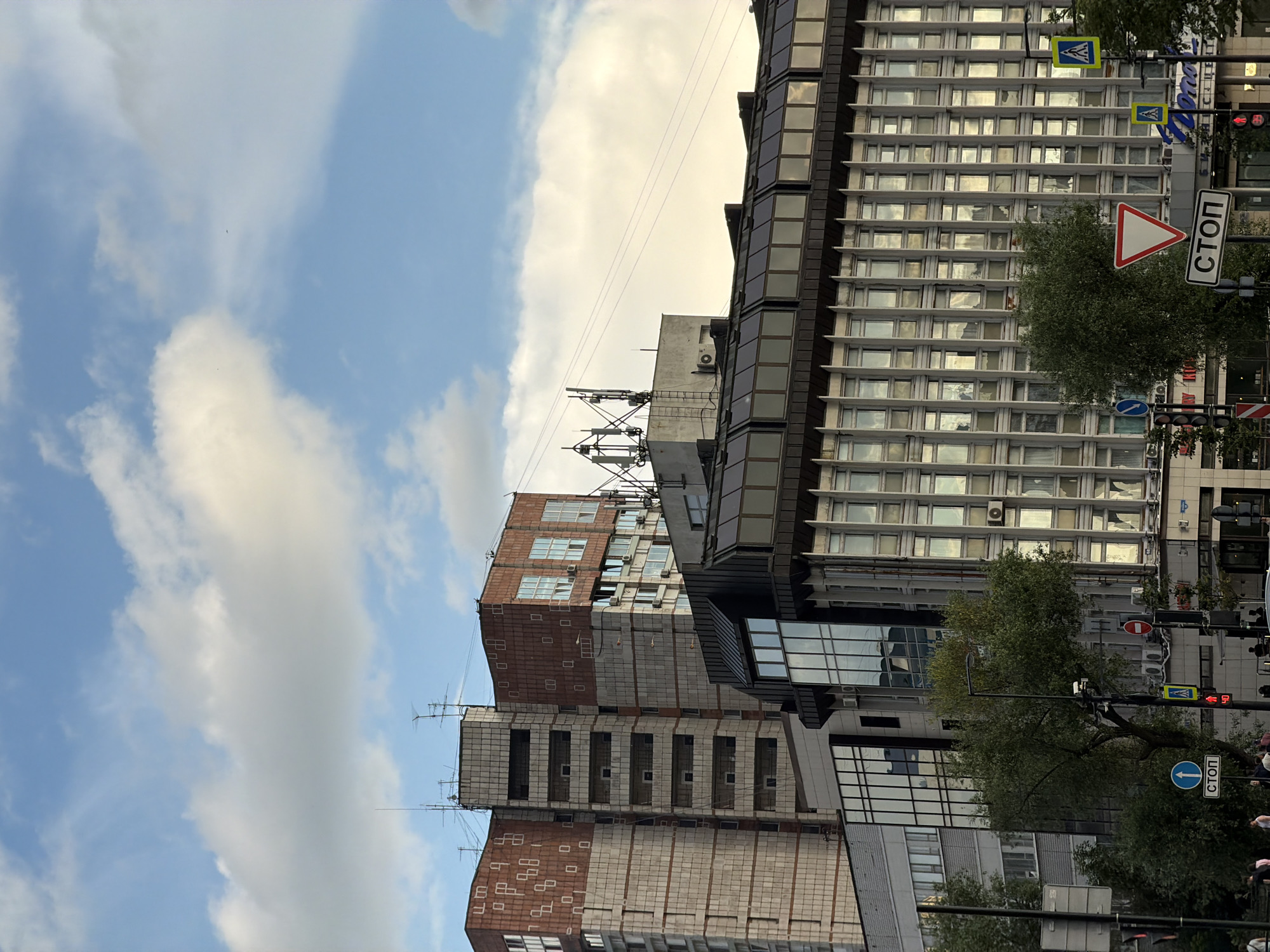 The image depicts a street scene in the city of Perm, Russia, with several buildings visible. In the foreground, there are traffic signs, including a stop sign and a pedestrian crossing sign. A traffic light is also visible. Trees line the street, and several people are walking in the background. The buildings are modern structures, some appearing to be office buildings or apartment complexes, with glass facades and various architectural details. One building has visible antennas on its roof. The sky is blue with scattered white clouds. The overall impression is a typical urban environment in Russia.