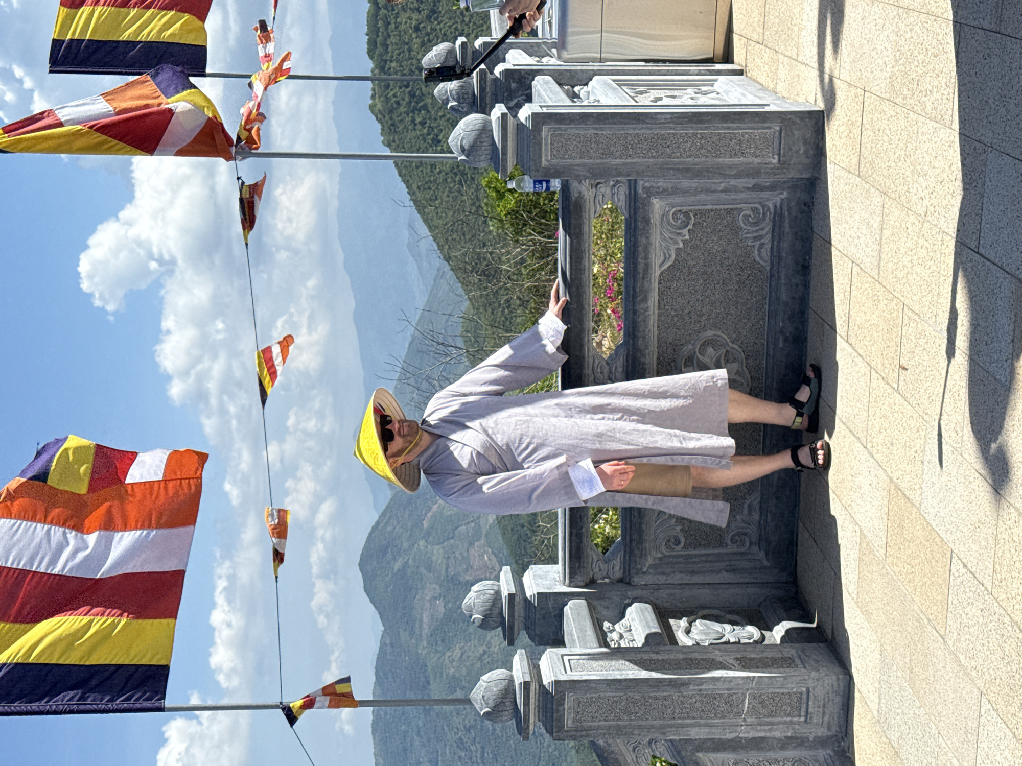 A tourist in a gray robe and yellow hat stands on a stone terrace in Nam Nha Trang Ward, Vietnam. The tourist holds a phone mounted on a selfie stick. A stone balustrade with carved lotus bud finials lines the terrace. In the background, distant mountains and a blue sky are visible. Several Buddhist flags are on poles in the foreground. The terrace floor is made of light-colored stone tiles.