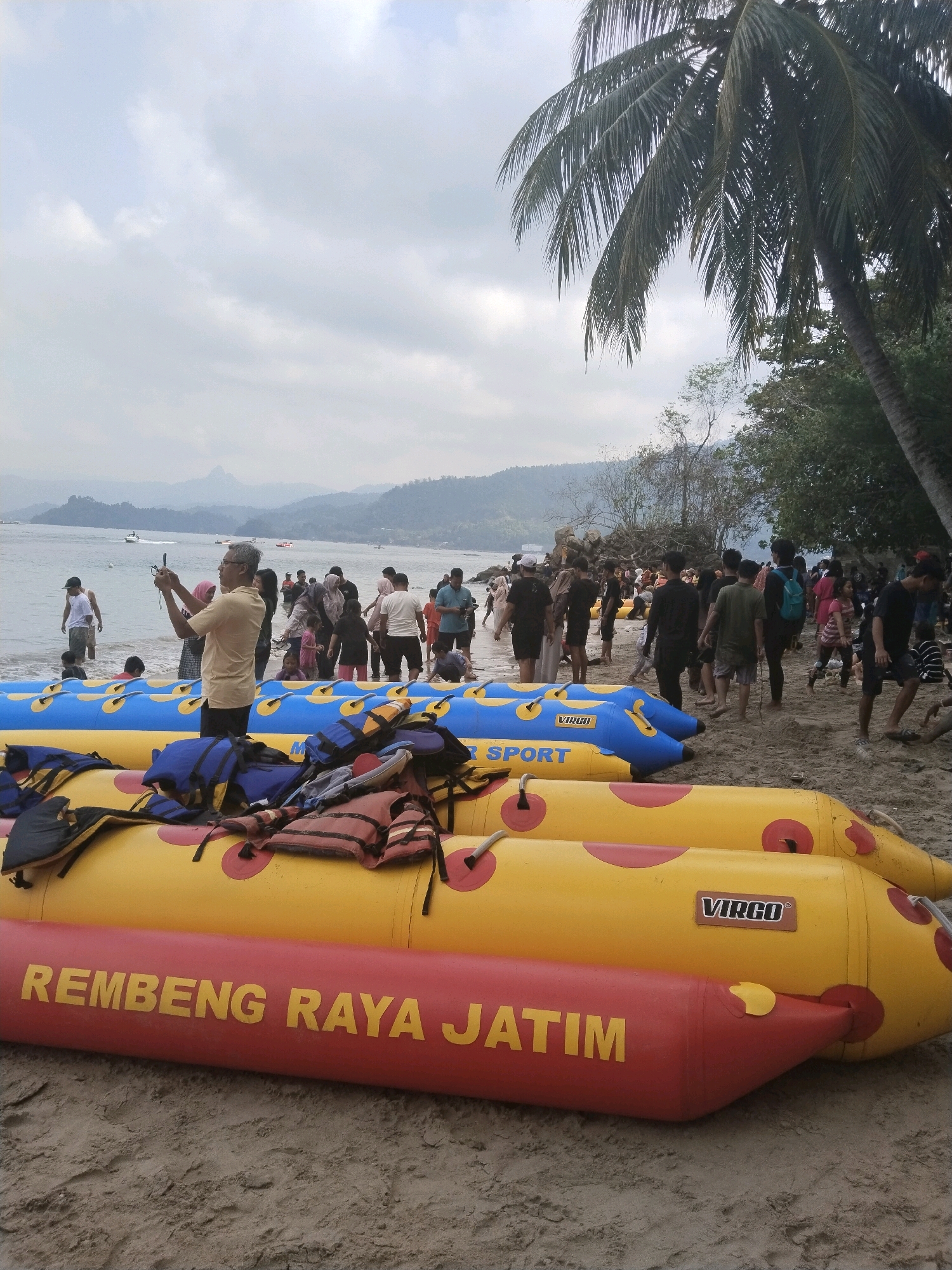 The image depicts a bustling beach scene in the city of Prigi, Indonesia, likely during daylight hours under an overcast sky. In the foreground, three inflatable banana boats are beached on the sand. The bottom boat is red with yellow lettering that reads "REMBENG RAYA JATIM". The middle boat is yellow with red polka dots and a label that says "VIRGO SPORT". The top boat is blue with yellow polka dots and also displays "VIRGO SPORT". Life vests, predominantly blue, red, and orange, are piled on top of the yellow and blue banana boats.
A crowd of people are visible on the beach and in the shallow water. Many appear to be tourists, dressed in casual beachwear. Some are walking along the shore, while others are standing around or wading in the sea. In the middle ground, a man in a yellow shirt is holding up a smartphone, seemingly taking a photograph or video. Behind him, a diverse group of individuals, including men, women, and children, are gathered. Further in the background, the sea stretches out, with a few boats visible on the water. The coastline is lined with hills covered in vegetation, and a hazy sky with clouds completes the scene. A tall palm tree is visible in the upper right portion of the image, suggesting a tropical or subtropical climate. The overall atmosphere appears to be one of leisure and recreation.