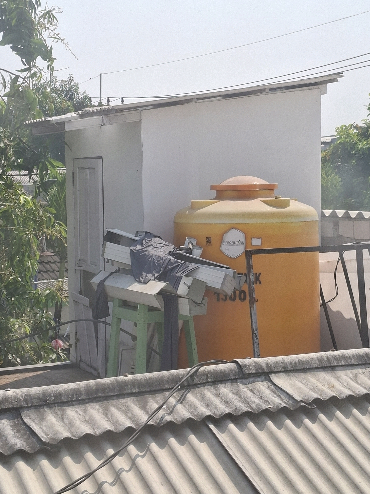 The image depicts a rooftop scene in Surabaya, Indonesia, under a clear, bright sky. In the foreground, a grey, corrugated roof is visible, with electrical wires running across it. Behind this, a white, rectangular structure stands, likely a small building or extension. To the right of this structure is a large, orange water tank with a lighter orange cap. The tank has some visible labels, including "SNI" and "PROFILTANK 1500 L". In front of the white structure, several long, metallic objects are propped up on a green stand, with dark fabric draped over them. To the left of the white structure, lush green foliage from trees, some bearing small green fruits, encroaches into the frame. The overall impression is one of an urban rooftop environment, possibly residential, with utilitarian elements like a water tank and storage.