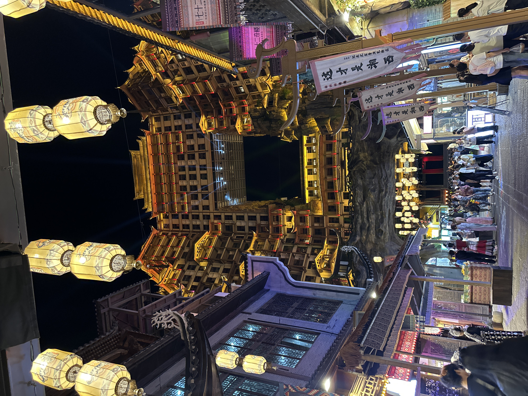 The image captures a vibrant night scene in Zhangjiajie, China, showcasing a traditional street lined with illuminated buildings designed in an ancient Chinese architectural style. Strings of lanterns cast a warm glow, highlighting intricate woodwork and ornate details of the structures. Several people are present, many of whom appear to be tourists observing the scene. One prominent building, constructed of wooden elements, rises multiple stories high with a central square opening. Street signs with Chinese characters hang overhead. The scene features a blend of historic architecture and modern tourism.