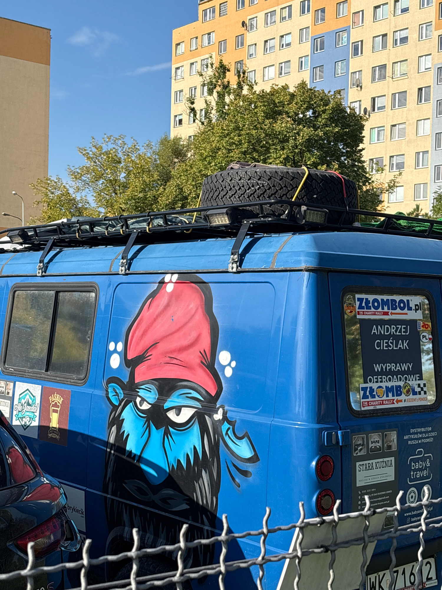 The image shows a blue van parked outdoors in Wrocław, Poland, in front of a large, colorful apartment building and trees. The van is equipped with a roof rack carrying a spare tire and several LED lights. A prominent graphic of a blue-faced character wearing a red hat is painted on the side of the van. Various stickers are affixed to the van's rear, including one that reads "ZŁOMBOL.pl THE CHARITY RALLY" and another with "ANDRZEJ CIEŚLAK WYPRAWY OFFROADOWE". Additional stickers indicate a business called "STARA KUŹNIA" which offers accommodation and "baby&travel". A portion of a black car is visible to the left of the van, and a wire fence runs across the foreground. The sky is clear and blue.