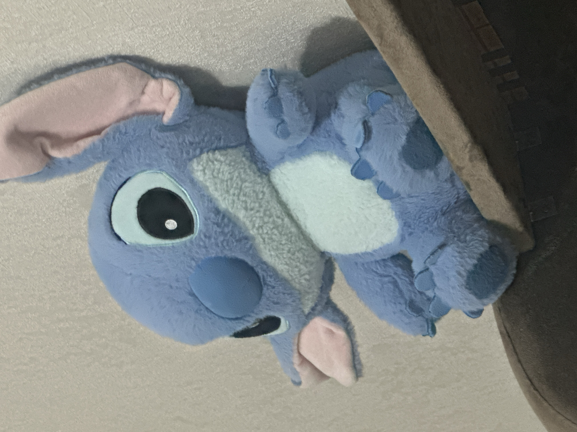 A blue plush toy resembling the character Stitch from Disney's Lilo & Stitch is positioned on a shelf against a textured light-colored wall. The toy is facing left with its head tilted slightly upwards. It has large, black eyes with white pupils, a light blue circular nose, and pink inner ears. Its mouth is slightly open, revealing a pink interior. The toy's body is predominantly blue with a lighter blue patch on its chest and blue paws with darker blue accents. It is sitting, with its arms resting on its lap. The shelf appears to be made of a brown, fabric-covered material. The background wall has a subtle, grainy texture. There is no identifiable location within the city of Chișinău, Moldova; the description is purely based on the visual content of the image.