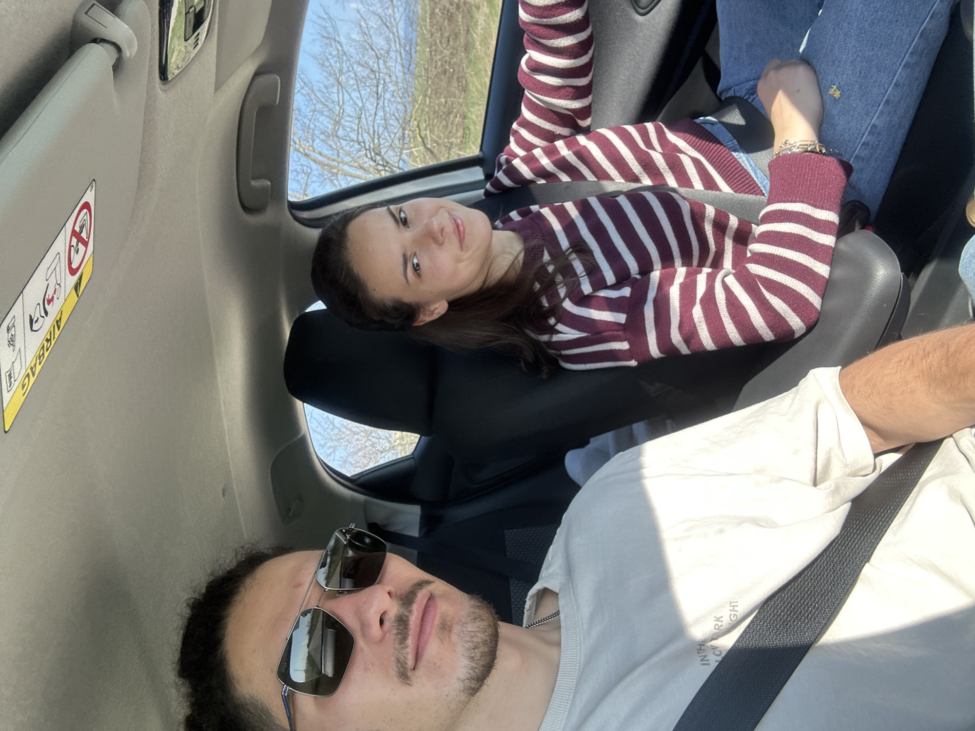 Inside a car, a man with sunglasses and a white t-shirt wearing a seatbelt is in the driver's seat. He is looking at the camera. A woman with a striped sweater and blue jeans is reclining in the back seat looking at the camera. The sky and trees are visible through the car windows. The location is not identifiable in this image.