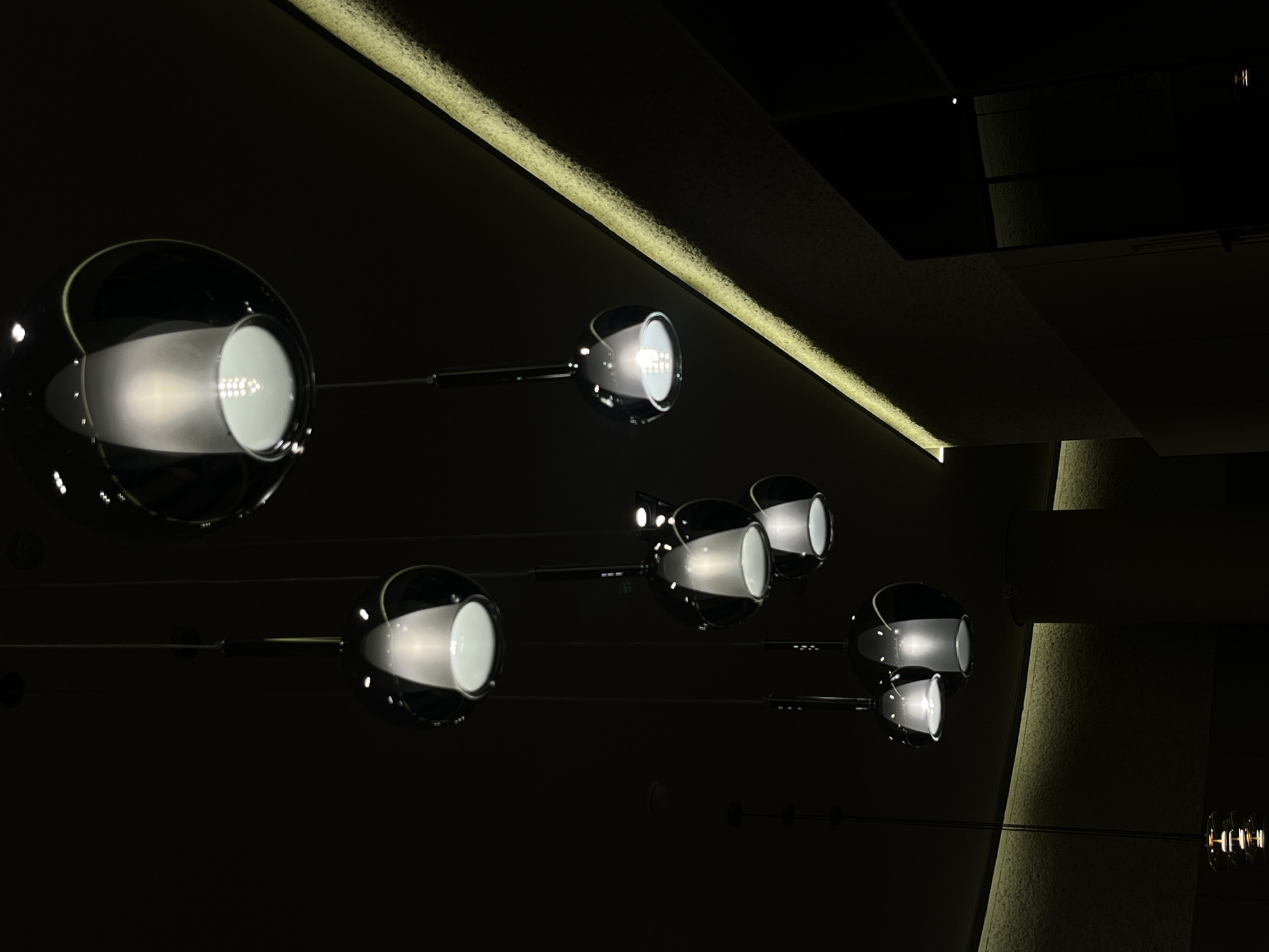 The image displays an interior scene, presumed to be in Mexico City, Mexico, under dim artificial illumination. Multiple modern pendant lights are suspended from a dark ceiling. Each light consists of a spherical, dark, reflective outer shell encompassing an inner frosted conical diffuser that emits cool white light from an LED array. These fixtures are arranged at varying heights throughout the frame. On the right, a textured vertical surface is illuminated by a horizontal strip of warm, indirect LED lighting, extending along its length. Dark, reflective paneling is visible in the lower right portion of the frame, showing faint reflections of light sources. A different, segmented cylindrical pendant light is partially visible at the bottom left. No individuals, animals, or specific actions are depicted.