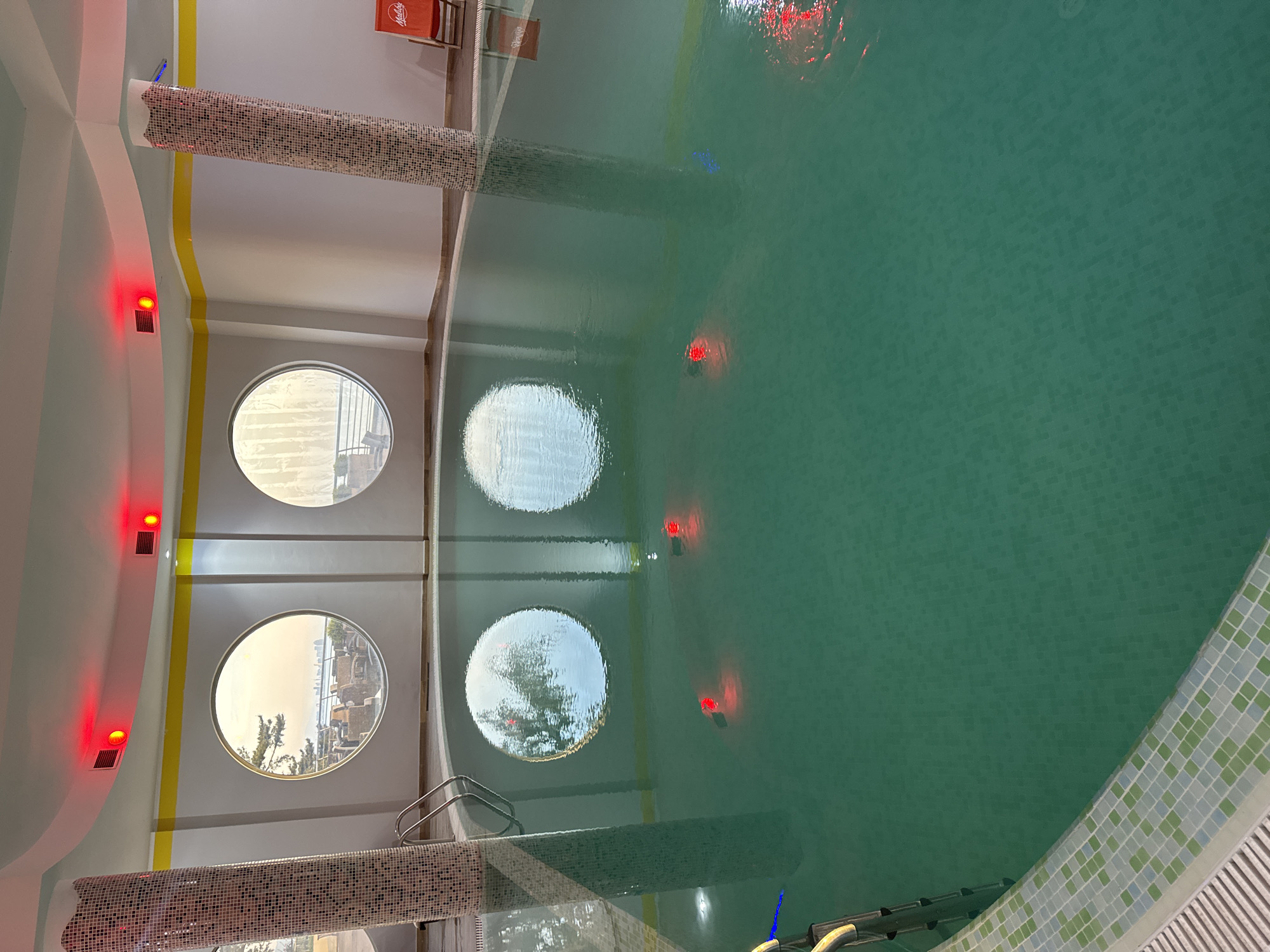 This image captures an indoor swimming pool area, viewed from an overhead perspective and rotated 90 degrees clockwise, placing the pool's length vertically. The pool water is a clear, deep teal color, and its bottom is tiled with small, uniform squares. Several red lights are submerged within the pool water, creating bright reflections on the surface, along with some fainter blue and white lights.
The pool is bordered on the right by a mosaic tile pattern of light green, white, and a pale grey. On the left side of the pool, a set of metal ladders extends into the water.
The surrounding architecture features light-colored walls with a yellow stripe running horizontally along the upper section. Two prominent round windows are set into the left wall, looking out onto a bright daytime scene with a clear sky and distant buildings or foliage. Reflections of these windows are visible on the pool's surface. Above these windows, two square red lights or vents are mounted on the wall.
Two decorative columns, covered in small mosaic tiles of white, brown, and grey, are positioned around the pool. One is prominently in the foreground on the left, and another is further back on the right. In the far background, outside the immediate pool area, red chairs with the "Maltesers" logo are visible, suggesting a commercial establishment like a hotel or resort. No people are present, and no specific activity is taking place; the scene is static. The overall impression is one of a well-lit, clean, and modern swimming facility.