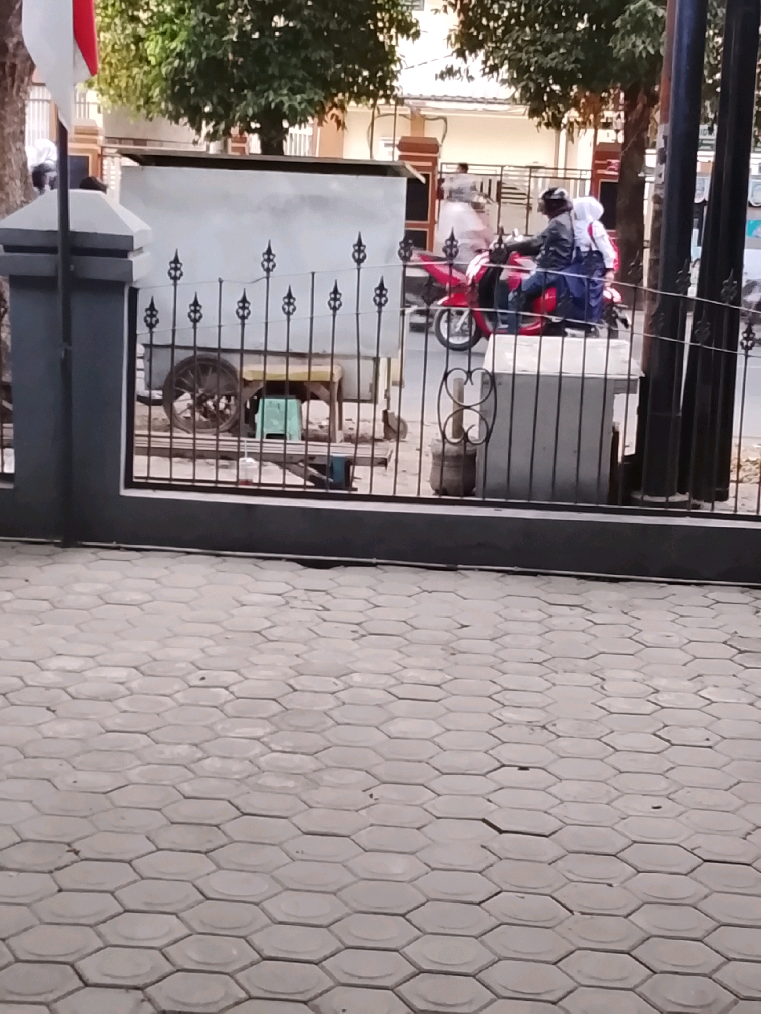 This image captures a street scene in Slawi, Indonesia, likely near a residential or commercial area given the presence of a gate and sidewalk. In the foreground, a patterned hexagonal paver sidewalk dominates the lower portion of the frame. Beyond this, a dark metal fence with decorative elements separates the sidewalk from the street. Through the fence, a red and white flag is visible on a flagpole to the left. A portion of a white building with a dark roof is also visible behind the fence.
In the middle ground, a red scooter with two occupants is traveling from left to right. The driver, wearing a dark jacket and helmet, is seated at the front, while a passenger, wearing a white hijab and a blue outfit, is seated behind. The scooter is positioned on a paved road, and a portion of a street cart with a large wooden wheel is visible behind the fence. The background features trees and buildings, typical of an urban Indonesian setting. The overall lighting suggests it is daytime.