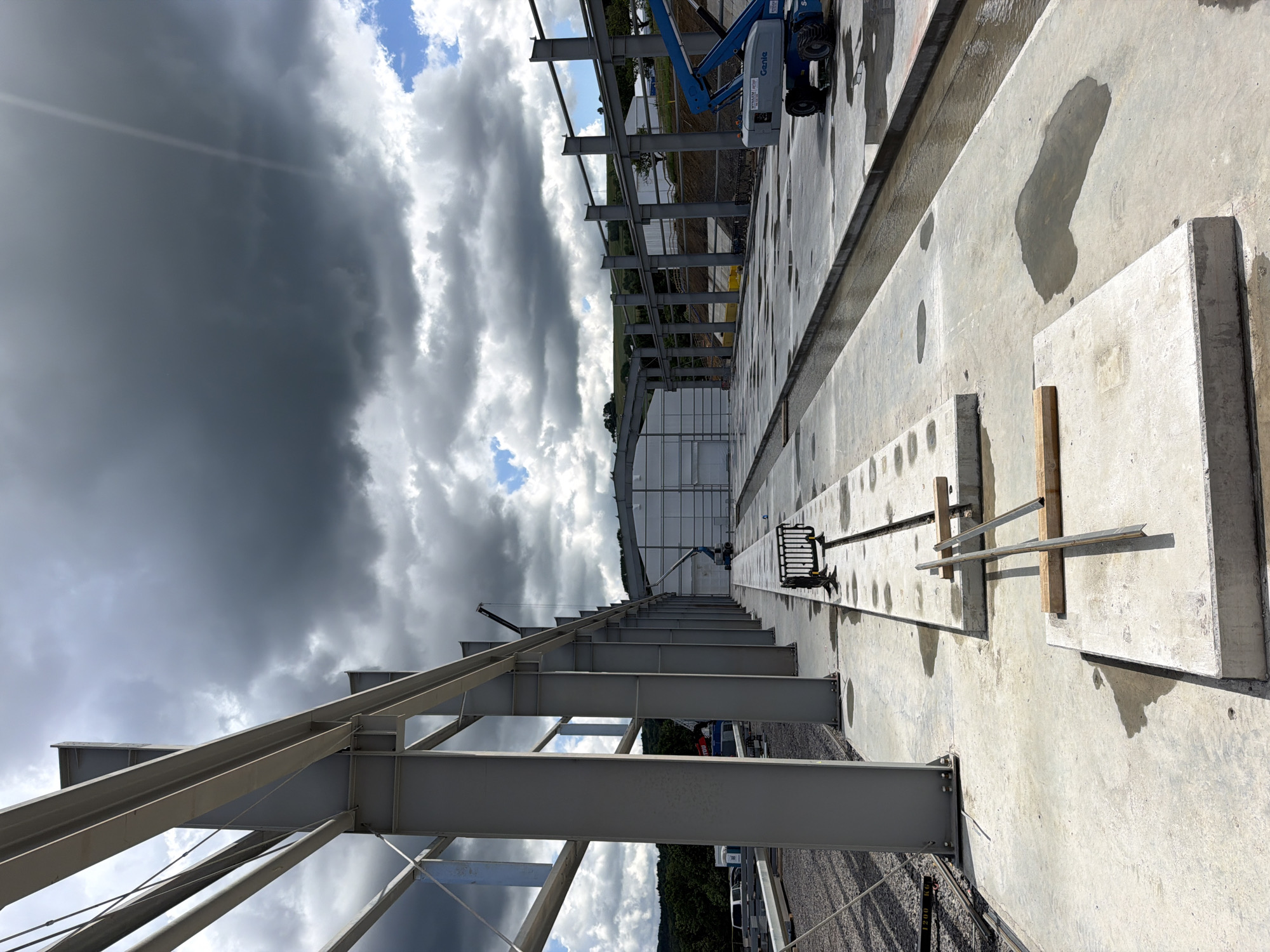 The image depicts an outdoor construction site in Stróża, Poland, characterized by unfinished steel structures and concrete foundations under a dramatic, cloudy sky. In the foreground, large gray steel beams are erected, supported by concrete pillars. The ground is a mix of concrete slabs and gravel, with some areas featuring puddles of water, suggesting recent rain. Several concrete elements are laid out, some with metal rebar protruding. A long, shallow channel of water runs alongside the constructed pathway. In the midground, a white building with a large door is visible, along with construction equipment including a blue boom lift (Genie brand) and cranes. The background shows rolling green hills and trees. There are no people clearly visible in the frame, but the presence of construction machinery indicates ongoing work.