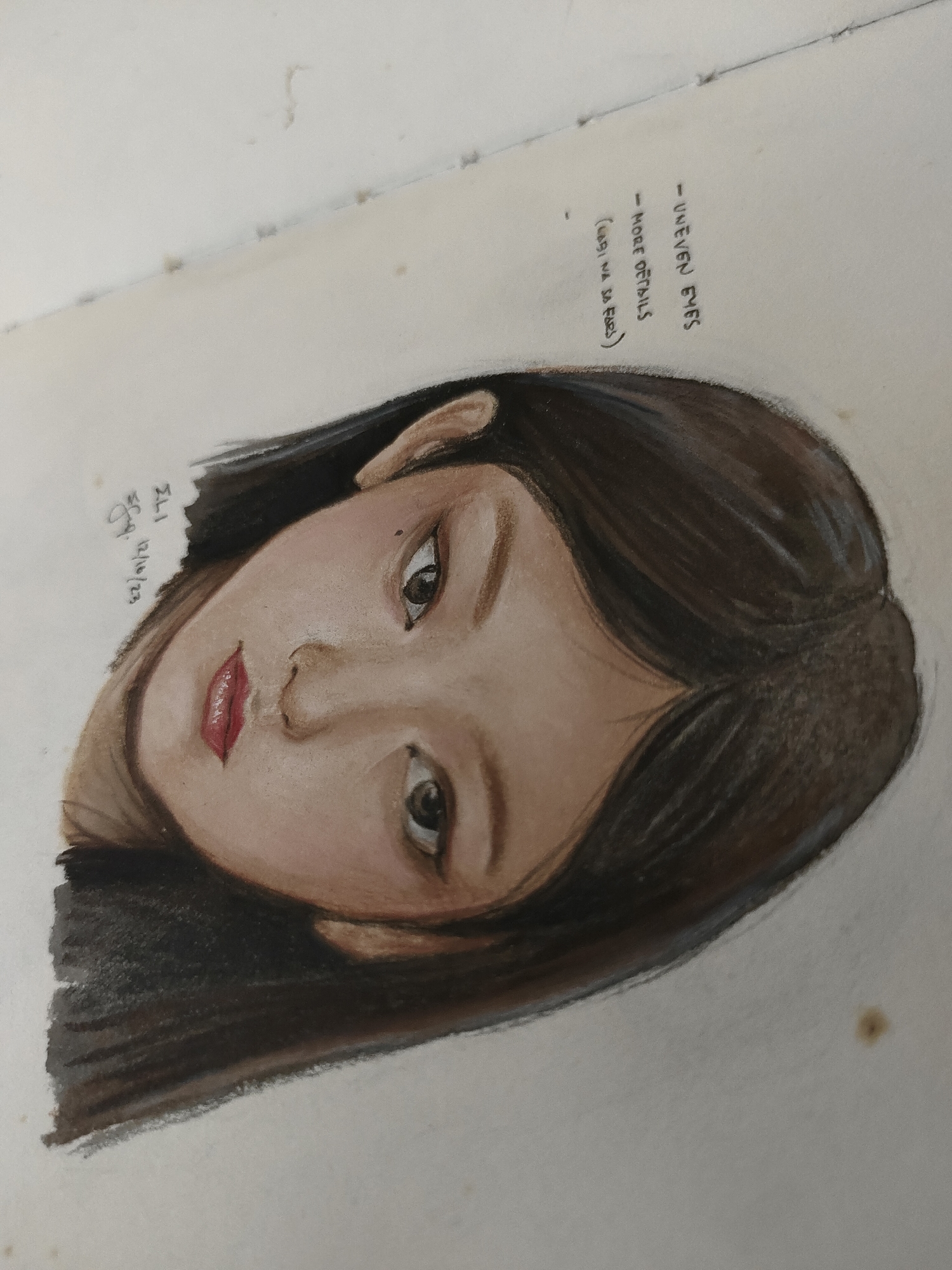 A top-down, slightly angled photograph captures a drawing of a female face on an off-white sheet of paper, rotated clockwise approximately 45 degrees. The drawing, executed with what appears to be colored pencils or pastels, depicts a young woman with dark brown hair framing her face and a light to medium skin tone. Her eyes are dark, gazing slightly downwards, with highlights indicating reflectivity. Her lips are a glossy red-pink, slightly parted.
Handwritten notes in black ink are visible on the upper right side of the paper, oriented vertically and reading upwards: "- UNEVEN EYES", "- MORE DETAILS", and "(LAGI NA BA AKO)". Below the drawing on the lower left, a signature "ELI Say" and the date "12/10/23" are written in similar black ink. The paper exhibits minor wear, including several faint brown spots and smudges. A faint horizontal line near the top edge suggests the paper may be from a sketchbook or notebook. The environment is an unidentifiable indoor setting in the city of Unknown, Unknown, indicated only by the clean, plain background against which the artwork is photographed.