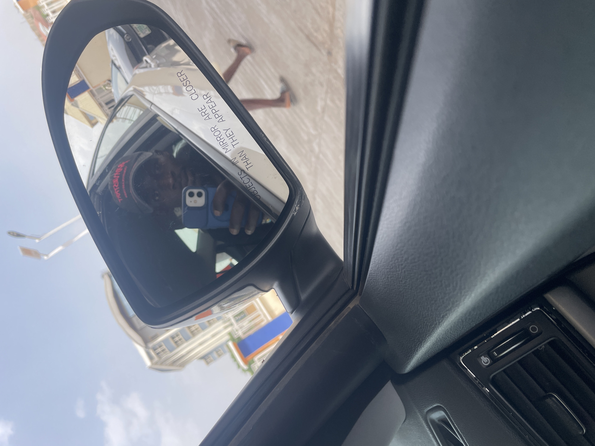 The image shows a side mirror of a vehicle, reflecting a person holding a phone, another vehicle, a building with blue and orange accents, and two legs of a person walking on the sidewalk. The mirror has the warning "OBJECTS IN MIRROR ARE CLOSER THAN THEY APPEAR." The interior of the car, specifically the air vent, is visible. The location appears to be Jos, Nigeria, based on the architectural style of the building and potentially the skin tone of the person reflected in the mirror.