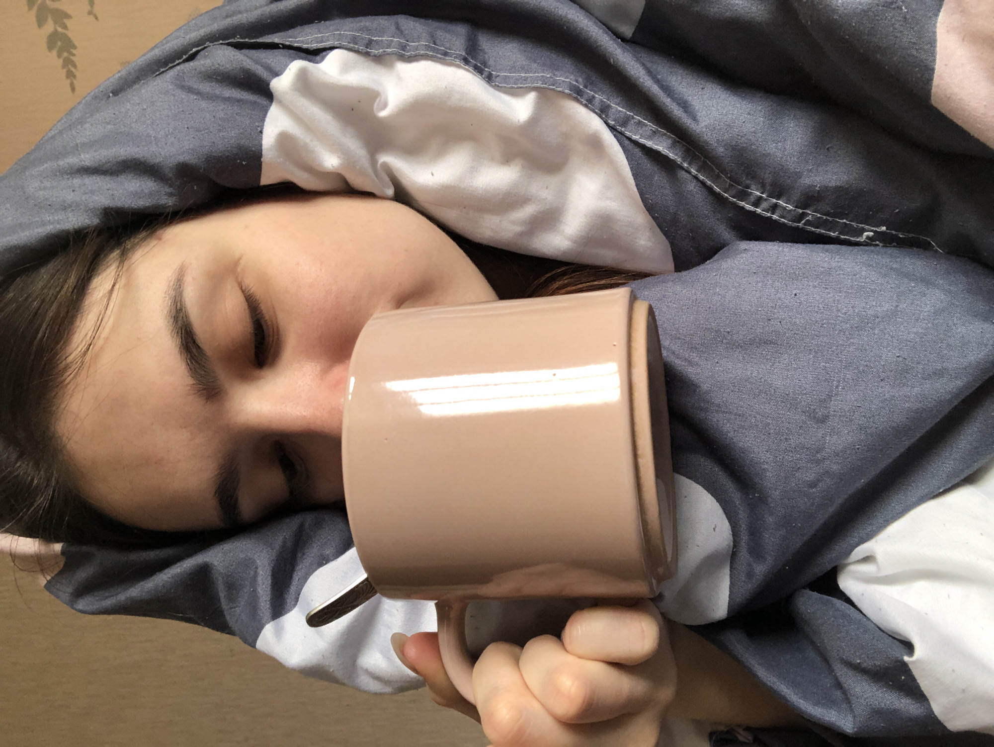 A person is lying in bed, under a gray and white patterned duvet. Their eyes are closed. They are holding a beige mug with a spoon in it. The background is a light tan wall. It is not possible to confirm the location is Chaikovsky, Russia, from the image.