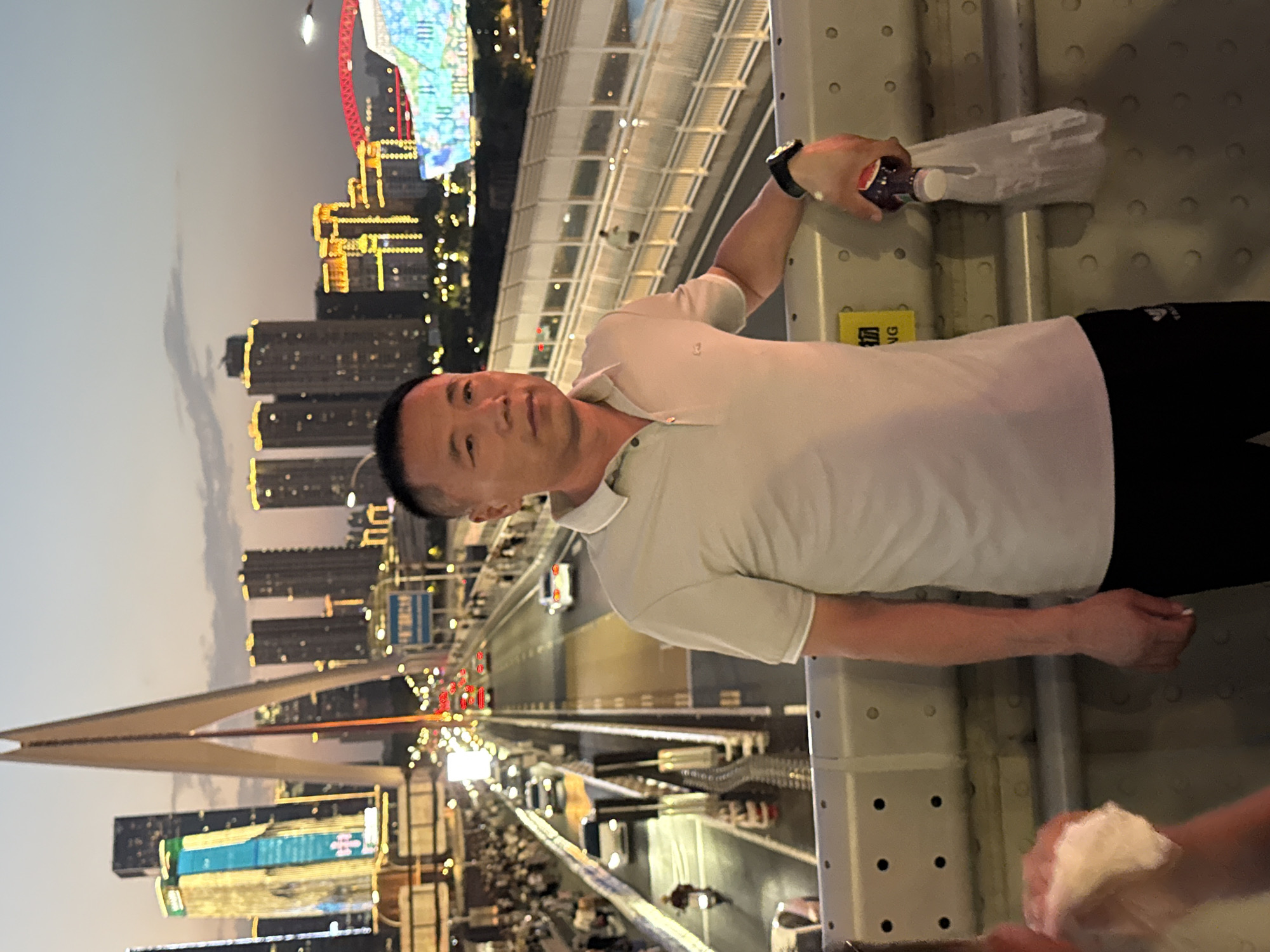 In Yuzhong District, China, at dusk, an Asian man stands on a bridge overlooking a busy cityscape. He is wearing a light gray polo shirt and black pants, with a watch on his left wrist. In his right hand, he holds a bottle of dark liquid and a plastic bag. The bridge has a metallic railing and a wall with circular indentations. In the background, skyscrapers illuminated with colorful lights rise against the twilight sky, and traffic flows on the bridge below. Pedestrians are visible on the bridge's walkways.