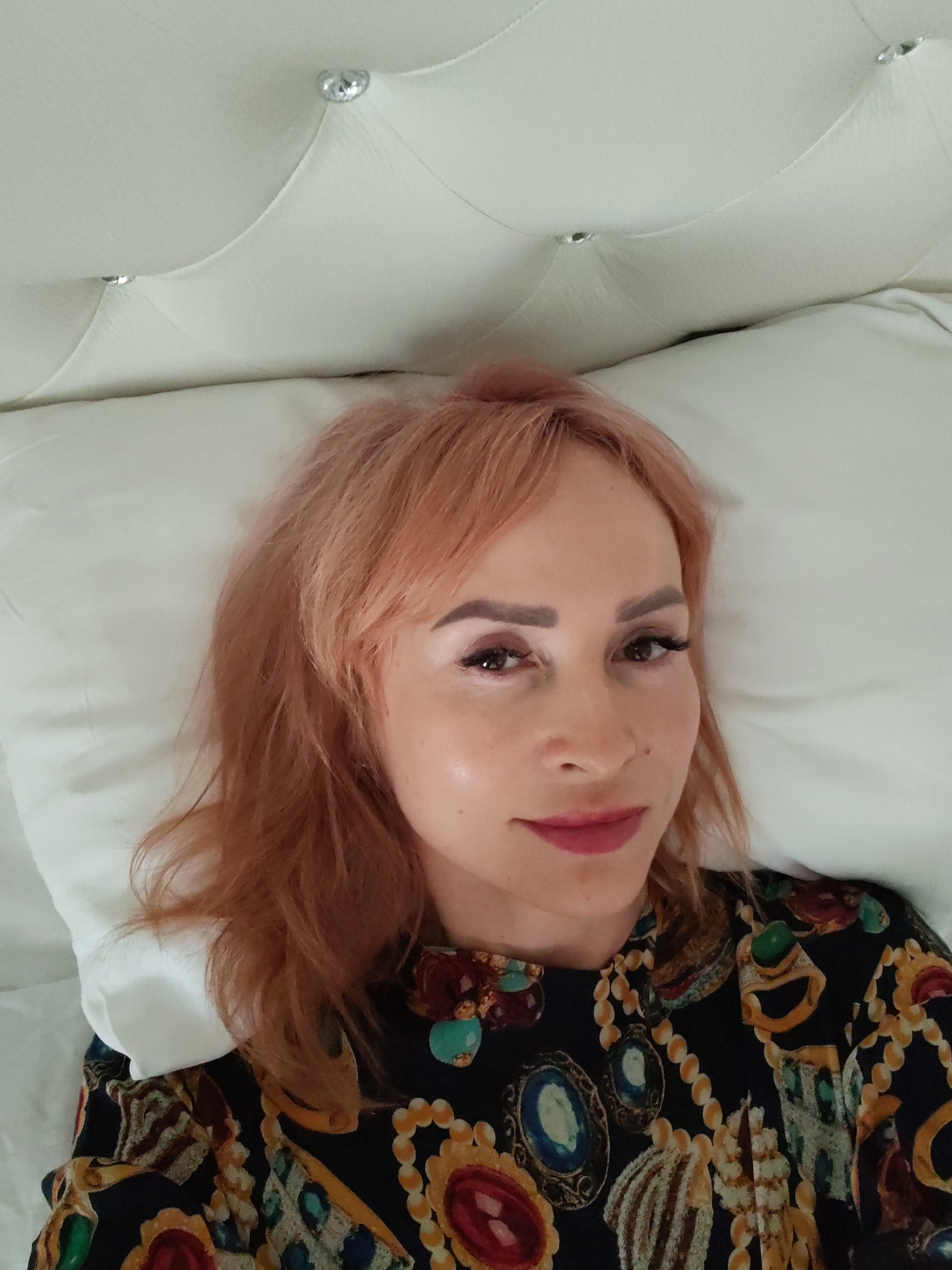 A medium shot, taken from a high angle, shows a woman with reddish-pink hair lying on a white pillow. She is looking up at the camera. Her eyes are open, and she is wearing makeup, including lipstick. She has on a dark-colored shirt with a colorful, ornate pattern featuring jewels and chains. The pillow is on a white tufted surface, possibly a bed, with decorative crystal accents on the tufting. The image is well-lit and appears to be taken indoors. The scene does not provide any specific visual cues to determine the location within the city of Nizhny Novgorod, Russia, or any other specific city.