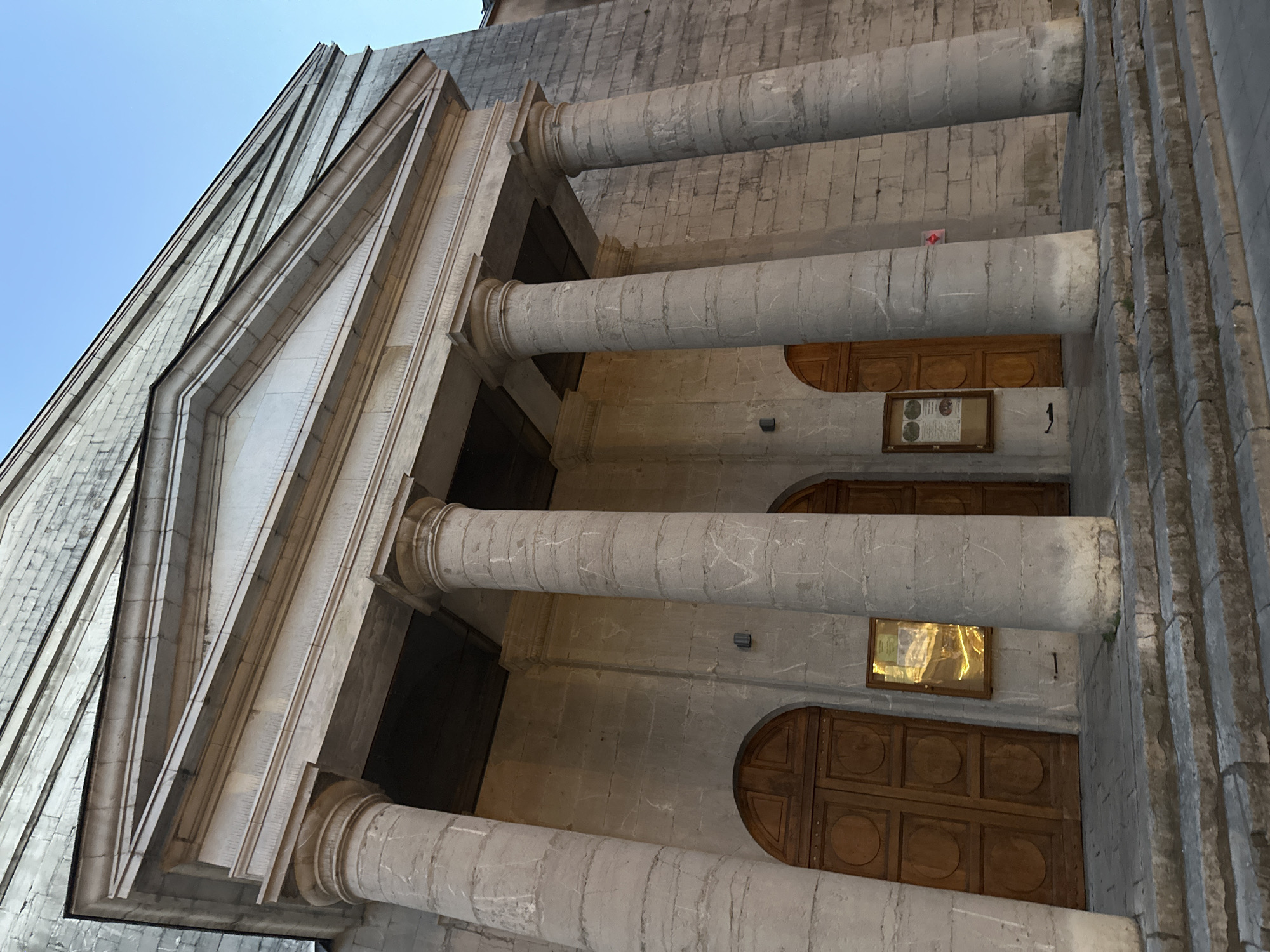 The image shows the facade of a stone building with classical architectural elements. There are several large, fluted stone columns supporting a triangular pediment. The entrance to the building appears to be through large wooden double doors, with at least two sets visible. There are also display cases mounted on the wall between the doors. The building is constructed from light-colored stone blocks, and the overall impression is one of grandeur and age. The sky above is a clear blue, suggesting daylight.
The building depicted is the Protestant temple in Anduze, France.