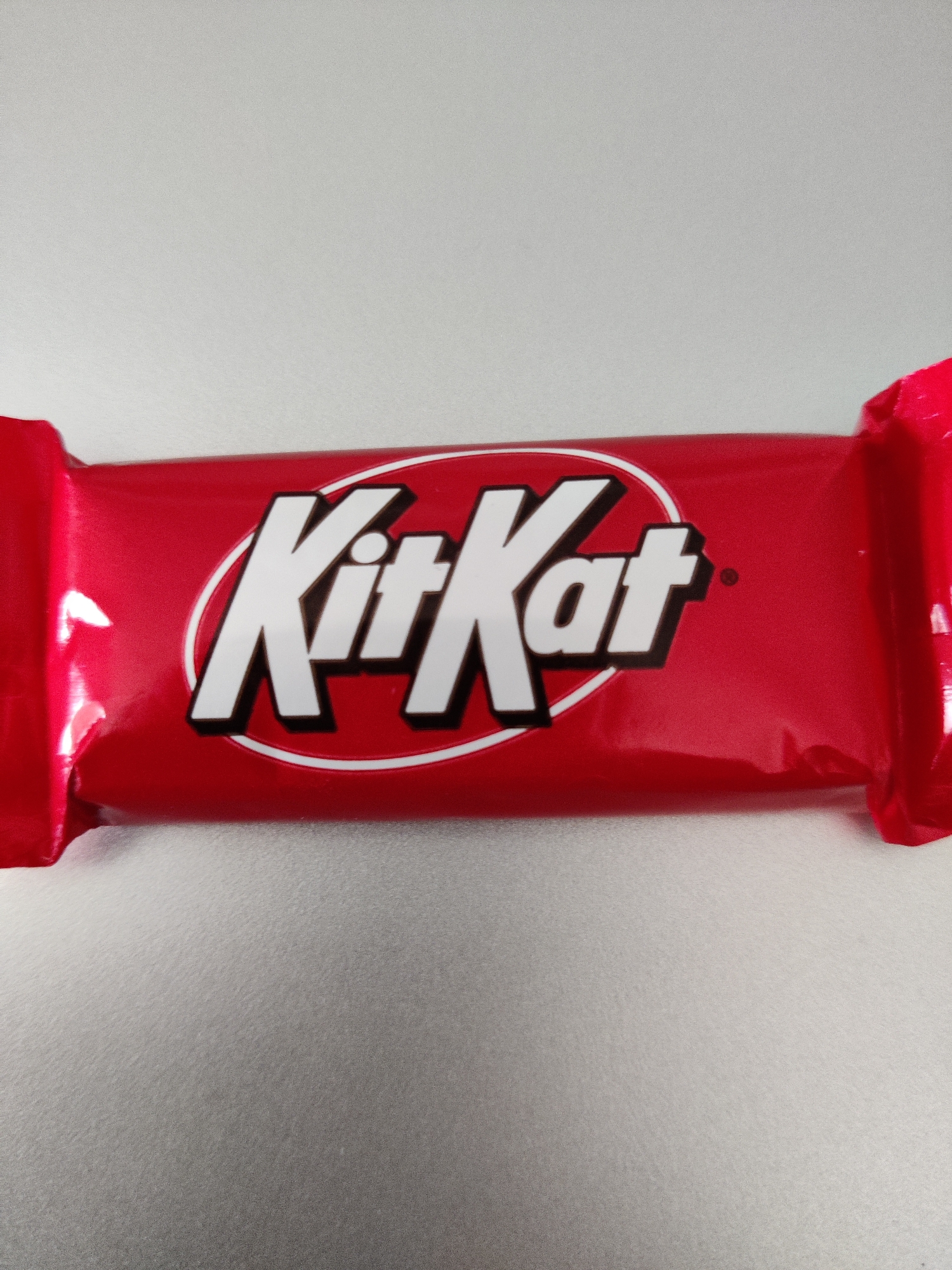 A Kit Kat candy bar wrapper lies horizontally on a textured grey surface. The wrapper is bright red with the white "Kit Kat" logo prominently displayed in the center, enclosed within an oval. The logo has a black outline and a slight shadow effect. The ends of the wrapper are twisted and folded, with the red material crinkling and reflecting light. The background is a plain, light grey.