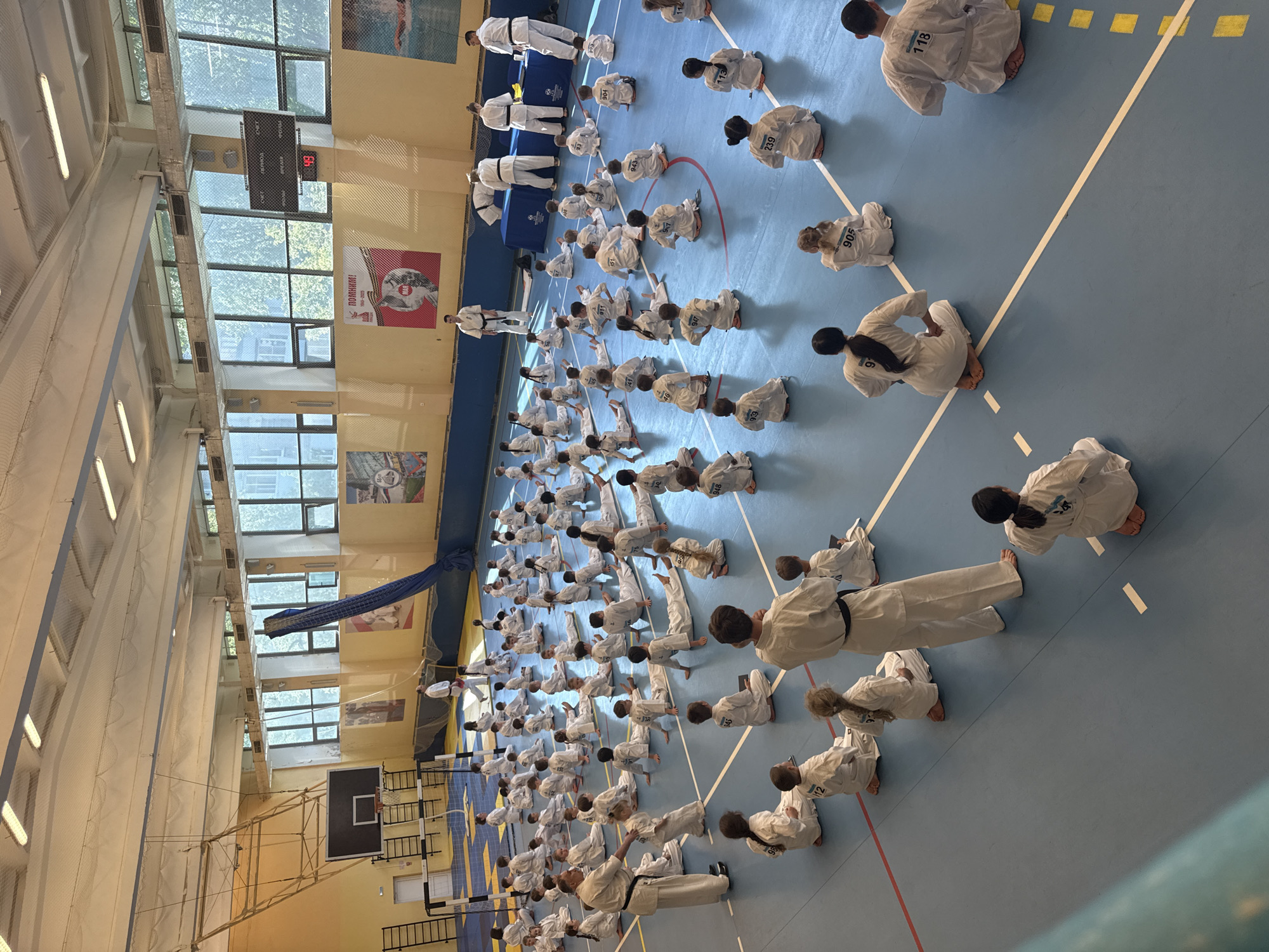 A large group of people are participating in a karate practice or competition inside a gym. The participants are primarily dressed in white karate uniforms and are arranged in rows on a blue floor marked with white and red lines. Some have numbers on their backs. Several people, possibly instructors or judges, wear black belts. The gym appears to be located in Yekaterinburg, Russia, as evidenced by signage in Russian. The gym has a basketball hoop and a scoreboard. Large windows let in natural light. Some people are standing while most are kneeling or sitting on the floor. The overall impression is a structured and focused martial arts event.