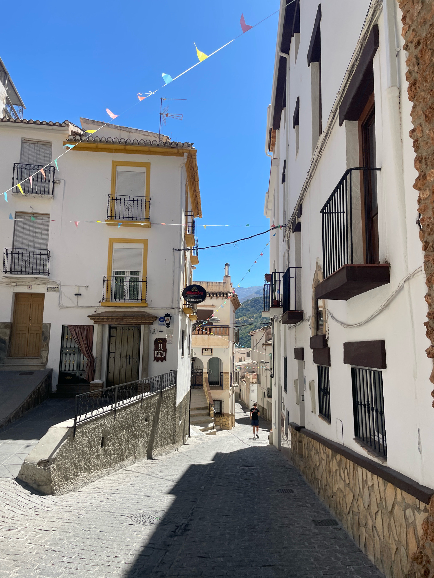 A man in a black t-shirt and shorts walks uphill on a narrow, cobblestone street in Güéjar Sierra, Spain. The street is flanked by white buildings with dark trim and balconies. Colorful triangular flags are strung across the street, and mountains are visible in the distance under a clear blue sky. One building features a sign for "El Portok" bar. The scene is brightly lit by the sun, casting distinct shadows on the street.