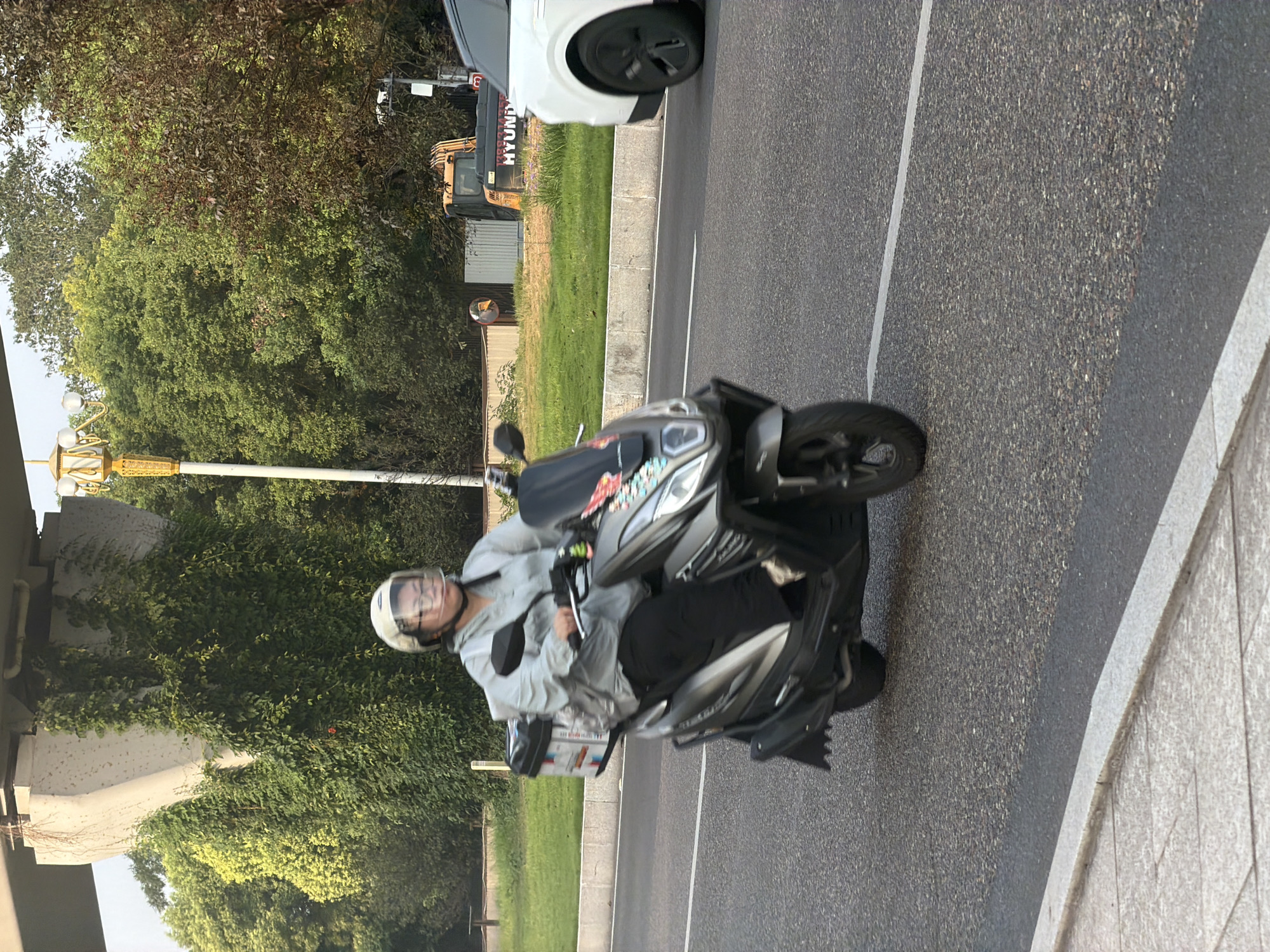 In the city of Jiangjin, China, a person wearing a helmet and glasses rides a gray and black scooter on an asphalt road. The scooter has a black storage box attached to the back, which displays colorful graphics. The rider is wearing a light gray hooded jacket and black pants. The road is bordered by a strip of grass and a concrete barrier. In the background, there are trees, a large concrete structure covered in ivy, a lamppost with multiple lights, and a yellow construction vehicle. To the right of the scooter, a white car is visible. The scene appears to be captured during the daytime, with soft lighting.