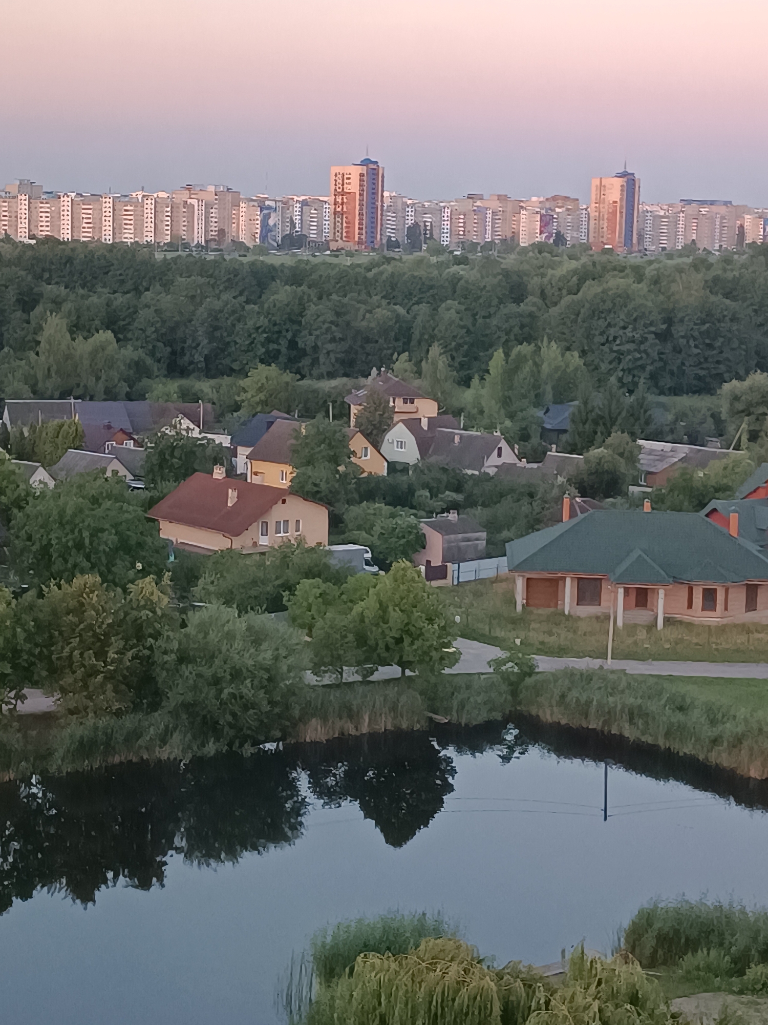The image depicts a panoramic view of a residential area with a body of water in the foreground, likely a pond or small lake. In the middle ground, a dense canopy of green trees separates the water from a cluster of houses, predominantly single-family homes with red or green tiled roofs. Some of these houses appear to be under construction, with visible brickwork and framing. Beyond the houses, a line of taller, multi-story apartment buildings forms the distant skyline. The sky above is a soft gradient of pink and pale blue, suggesting either sunrise or sunset. The reflection of trees and houses is visible on the calm surface of the water. The scene is devoid of any visible people or vehicles, contributing to a peaceful, suburban atmosphere. The overall setting suggests a developed urban or semi-urban area bordering a natural water feature and a wooded zone.