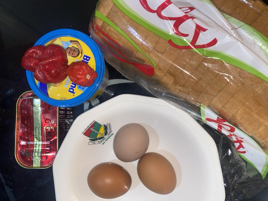 An overhead view in Tudun Nupawa, Nigeria, displays a collection of food items arranged on a dark, reflective surface. A white octagonal bowl, featuring a logo inscribed with "ALL LIVES ARE PRECIOUS" and "44 NARHK KADUNA," contains three brown-shelled eggs. To the upper left, a red and gold rectangular can of "OGBONGE" brand sardines in olive oil is visible. Adjacent to the right, a blue-lidded tub of "Blue Band" margarine or spread, depicting a child's face on its lid, is topped with two red peppers, one large and one smaller, possibly scotch bonnets. A clear plastic bag containing a sliced loaf of white bread with a red and green label partially visible occupies the lower right portion of the frame. No individuals are present, nor are any actions or interactions depicted.