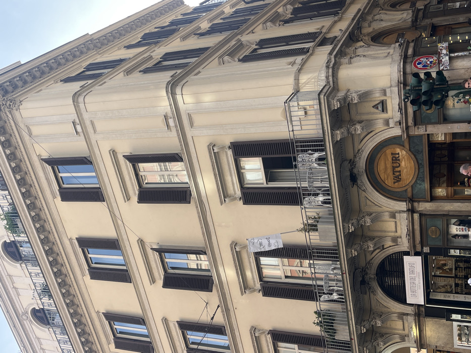 The image displays a multi-story, cream-colored building situated at the intersection of Via Condotti and Largo Carlo Goldoni in Rome, Italy. The building features classical architectural details, including decorative cornices and pilasters. Five floors are visible, each containing multiple rectangular windows fitted with dark louvered shutters, many of which are open, revealing reflections of the sky or interior spaces.
A metal-railed balcony extends along the second floor and wraps around the corner. On this balcony, two white ornate metal tables and four matching chairs are arranged. Several potted plants are visible in rectangular planters. A white rectangular flag or banner, displaying text including "cloud", hangs vertically from a pole attached to the wall above the balcony.
The ground floor hosts commercial establishments. On the left side of the main facade, a shop identified as "LA BOTTEGA DEL TARTUFO" is visible. At the building's corner, a prominent circular wooden sign reads "VATURI", positioned above an arched entrance. Below the "Vaturi" sign, a green traffic light pole with multiple signals and a circular red "No Entry" sign are present.
Two individuals are partially visible at the bottom right of the frame, near the traffic light. The scene is illuminated by natural light, with a clear blue sky visible at the top.