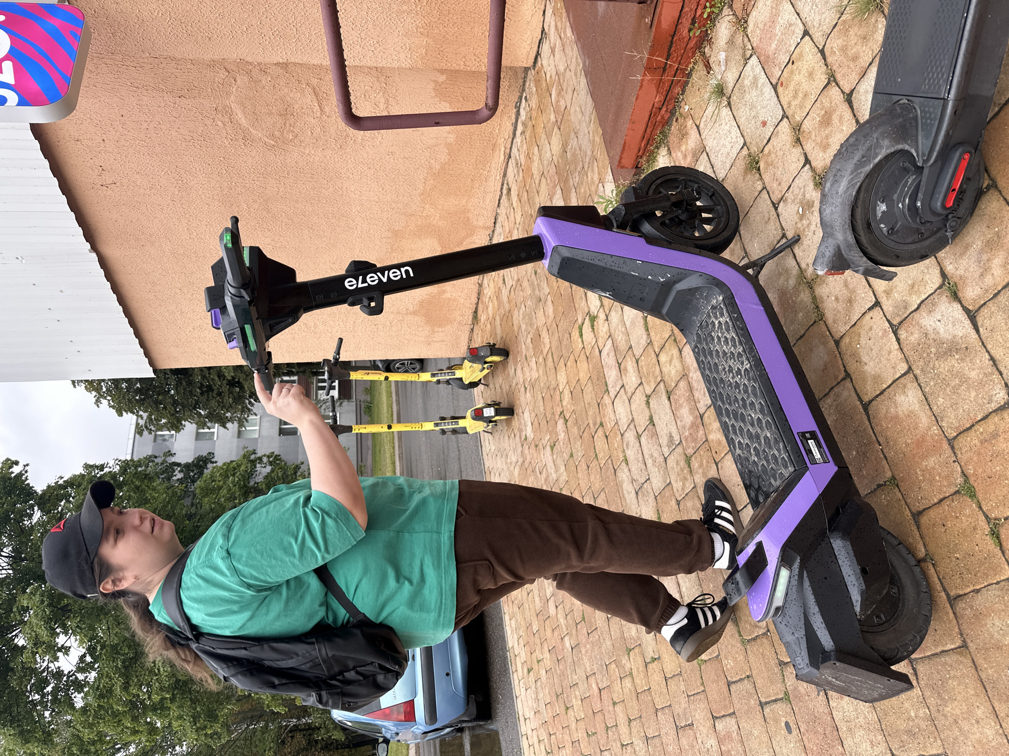 An individual wearing a green t-shirt, brown pants, black sneakers with white stripes, and a black baseball cap is standing on a paved walkway made of reddish-brown bricks. They are wearing a black backpack and are interacting with the handlebars of a black and purple electric scooter. The scooter has the "e-leven" logo on its stem and green indicator lights. Behind the individual and to their left, a blue car is parked on the street. Two yellow electric scooters are visible in the background, parked in a row. To the right of the main subject, a pink and blue neon sign is attached to an orange-colored building. The ground is wet, suggesting it has recently rained. The overall scene appears to be an urban environment, possibly a street or public area in Minsk, Belarus.