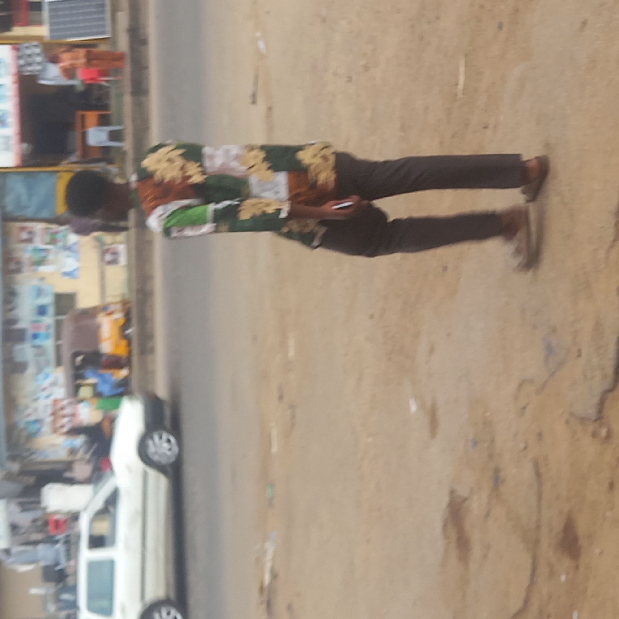 A man stands on a dirt shoulder next to a paved road in Sardauna, Nigeria. He is wearing a patterned, long-sleeved shirt, dark pants, and brown shoes. He is holding a cell phone. A white car is parked on the side of the road. In the background, there is a building with a solar panel on top, and what appears to be a small kiosk with various items displayed. Other vehicles and people are further in the background. The ground is dry and dusty.