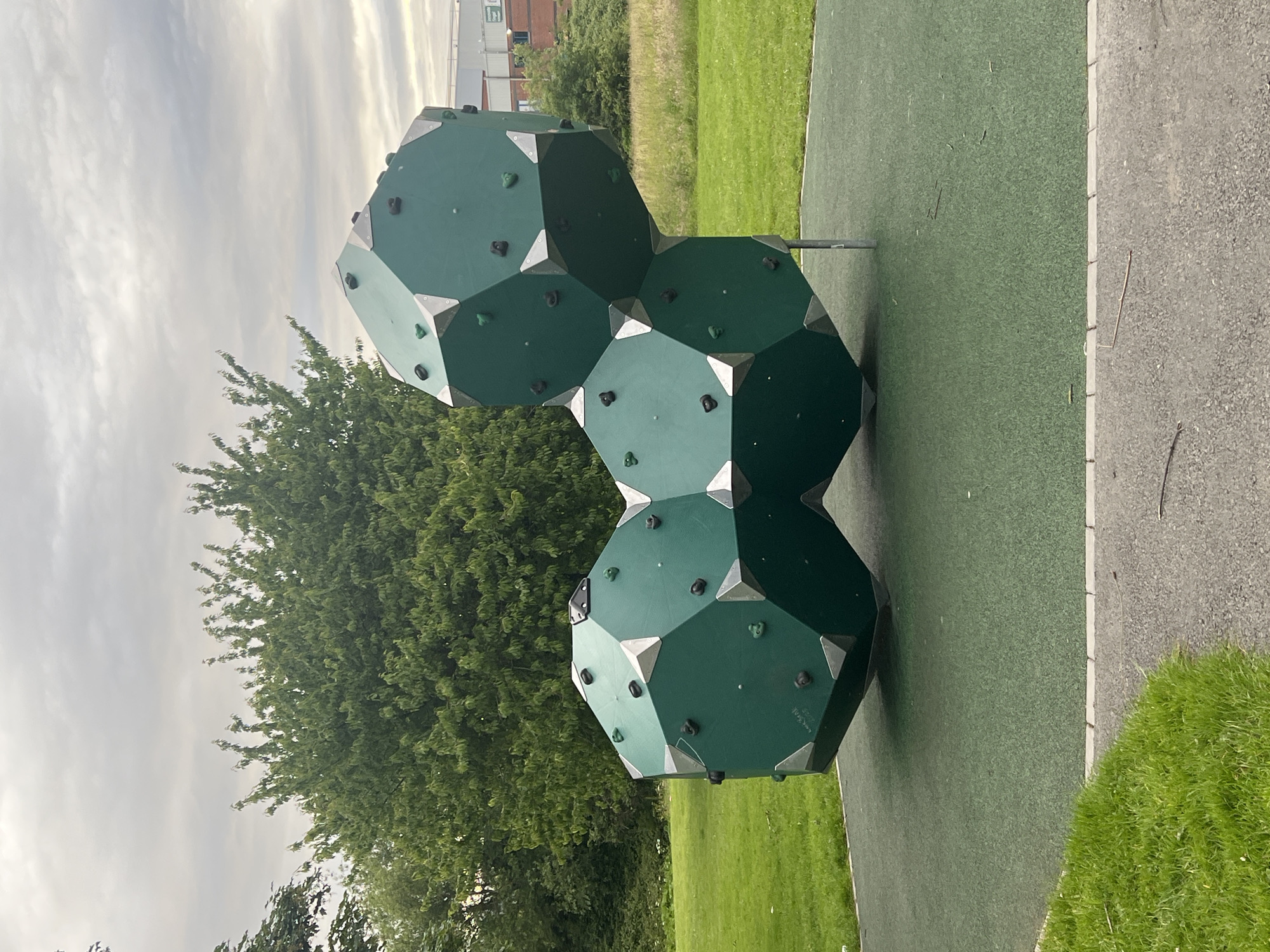 A cluster of geometric, dark green climbing structures, resembling connected octagons, is situated on a bright green rubberized play surface in a park. The structures are adorned with numerous black and some green climbing holds, as well as silver metal corner brackets. The play area is bordered by a light gray paved path and a strip of grass. In the background, a dense green tree line separates the park from a light-colored building with red accents and windows, indicative of an industrial or commercial establishment. The sky above is overcast with gray clouds. This scene is located in Balbriggan, Ireland.