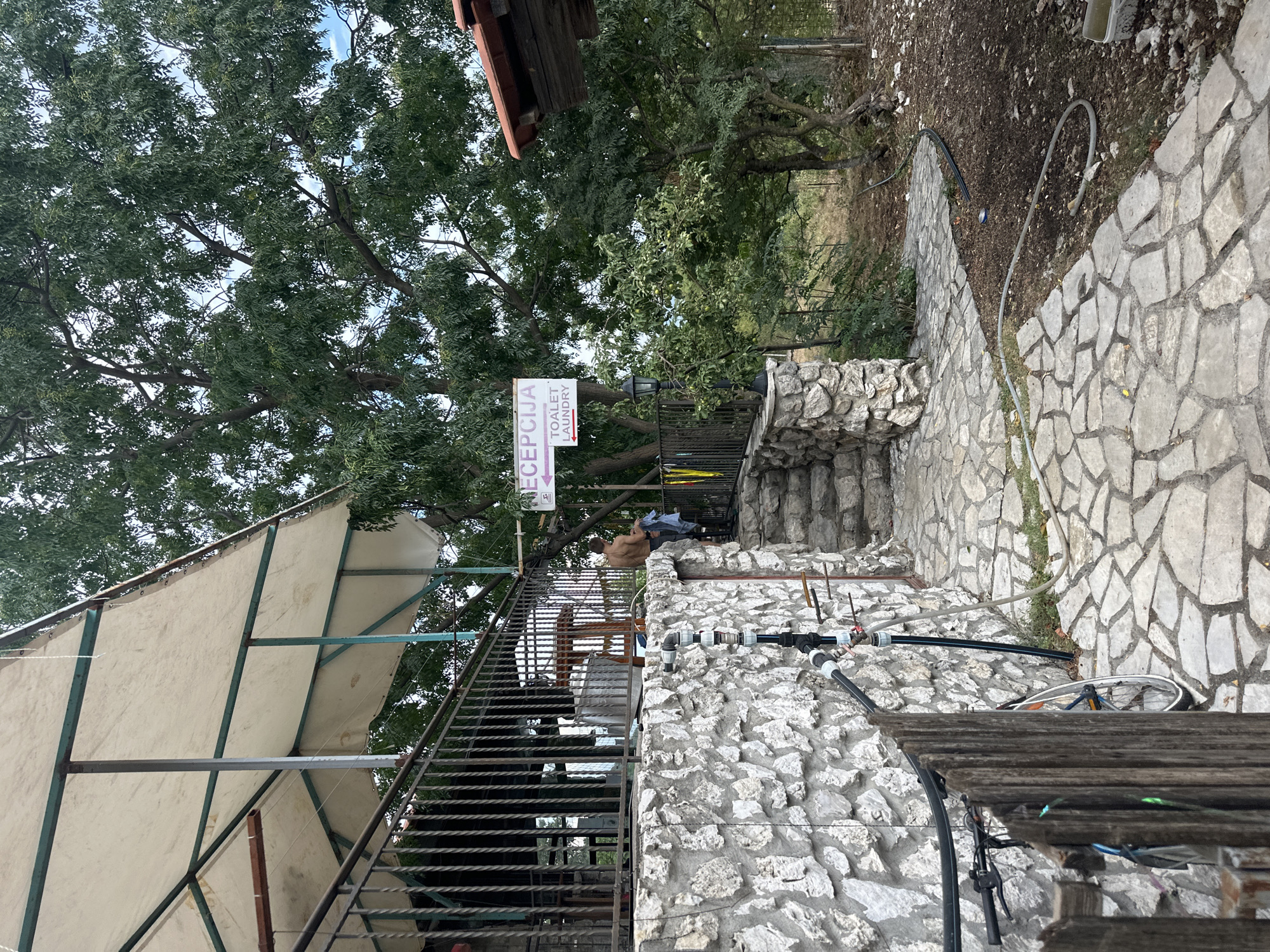 The image depicts an outdoor scene, possibly a campsite or a guesthouse, in Cetinje, Montenegro. A stone-built wall forms the foreground on the left, with a wooden fence and a bicycle leaning against it. A paved pathway made of irregular stone slabs leads towards a set of stairs. To the right of the stairs, a signpost indicates "RECEPTION" and "TOALET LAUNDRY" with arrows. A person is standing near the stairs, partially visible from behind, holding a blue garment. Above, a large tree with dense green foliage dominates the upper right portion of the frame, with a white canopy structure extending from the left side of the image. The overall environment appears rustic and natural, with trees and vegetation surrounding the built structures.