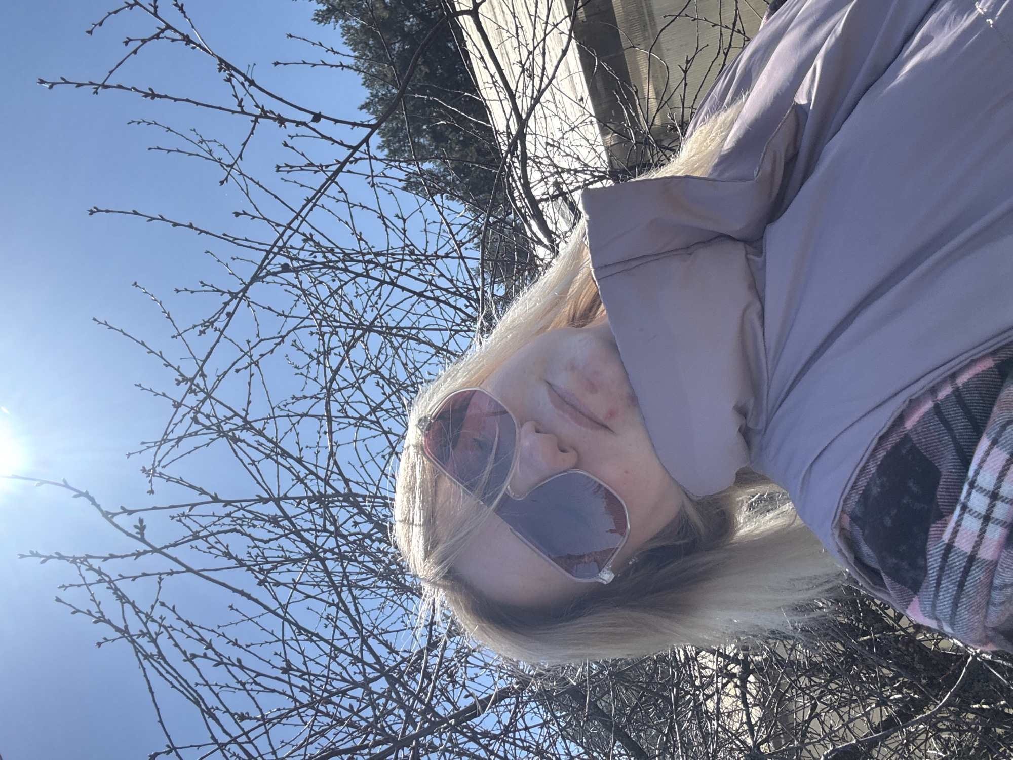 A low-angle, medium shot depicts a blonde woman wearing pink-tinted sunglasses and a pink puffer jacket with a plaid collar. She is positioned against a backdrop of bare tree branches silhouetted against a bright blue sky with the sun shining. Part of a concrete structure is visible in the background. The location is Chaikovsky, Russia.