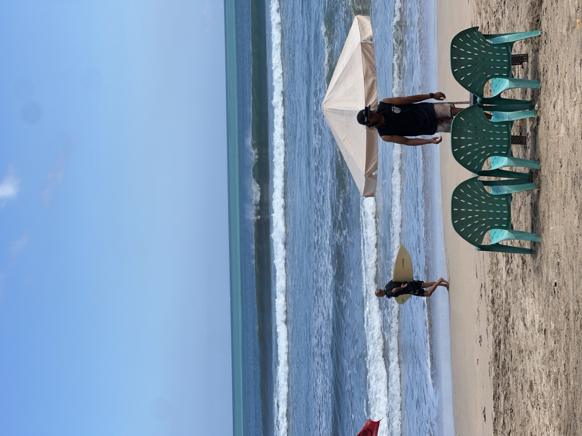 This is a vertical, medium-shot, eye-level, natural light photograph taken on a beach in Kuta, Indonesia. In the foreground, three green plastic chairs are arranged in a row on the sand. Behind the chairs, a large, light-colored umbrella is partially submerged in the ocean, with a man wearing a black sleeveless shirt and patterned shorts standing in front of it. Further back on the sandy shore, a surfer carrying a light-colored surfboard walks towards the water. A red flag is visible to the left of the surfer. The ocean in the background features several lines of waves, with the horizon showing a clear distinction between blue and turquoise water. The sky is clear and blue with a few faint clouds.