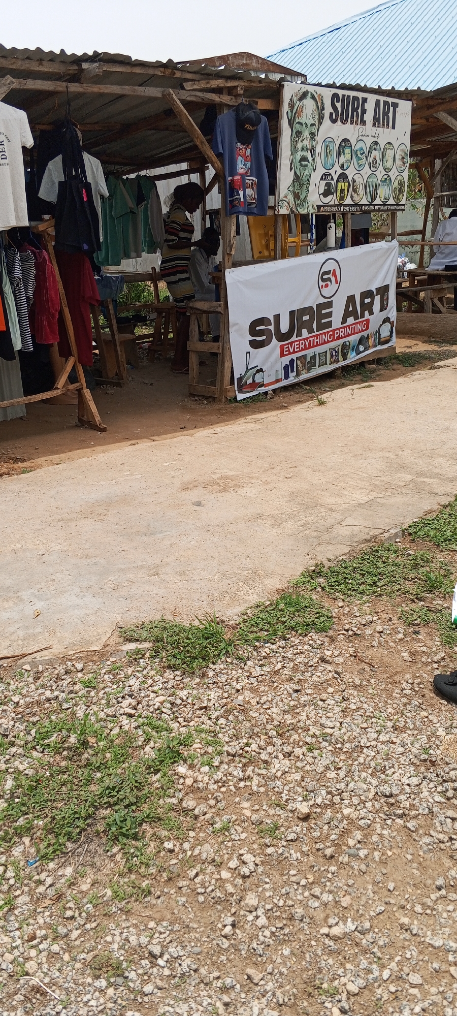 The image shows a roadside stall in Keffi, Nigeria. The stall is constructed of wood and corrugated metal, displaying clothing and artwork for sale. A sign prominently reads "SURE ART EVERYTHING PRINTING". Two people are inside the stall, potentially vendors or customers. The area in front of the stall transitions from a concrete surface to a gravel and grassy area.