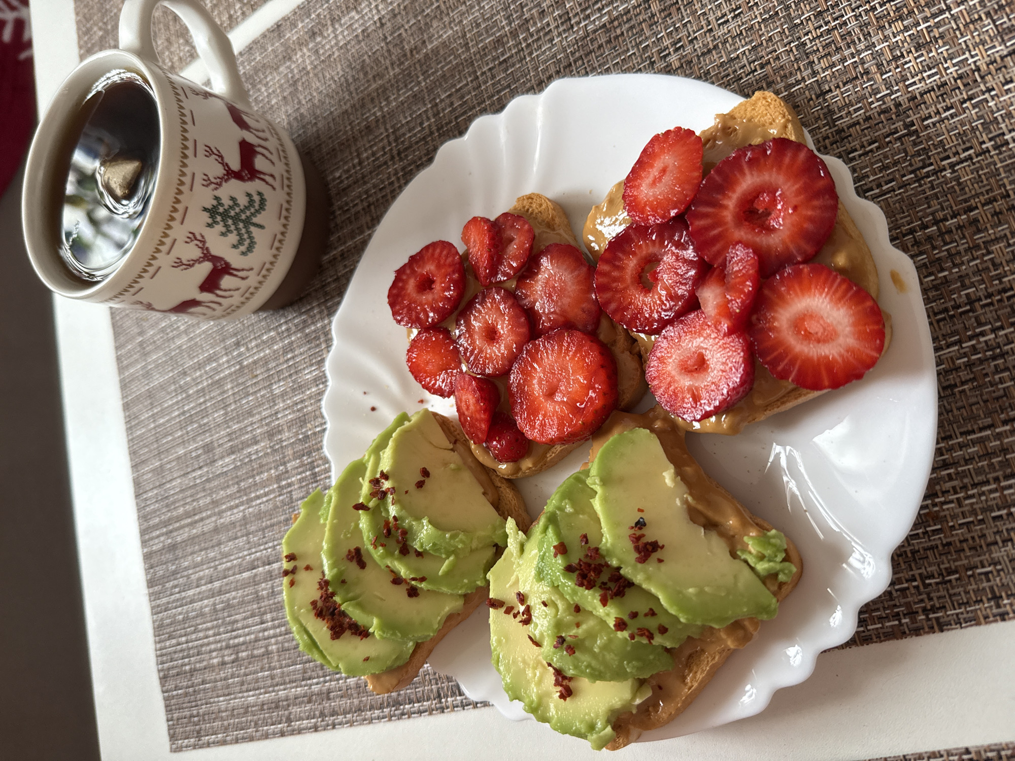 A close-up, top-down shot shows a white plate with four pieces of toast topped with peanut butter. Two of the toasts are topped with sliced avocado and red pepper flakes, and the other two are topped with sliced strawberries. To the right of the plate is a white mug with a Christmas design of reindeer and trees, filled with dark liquid and a tea bag. The plate and mug rest on a brown woven placemat on a white table. The background is blurred.