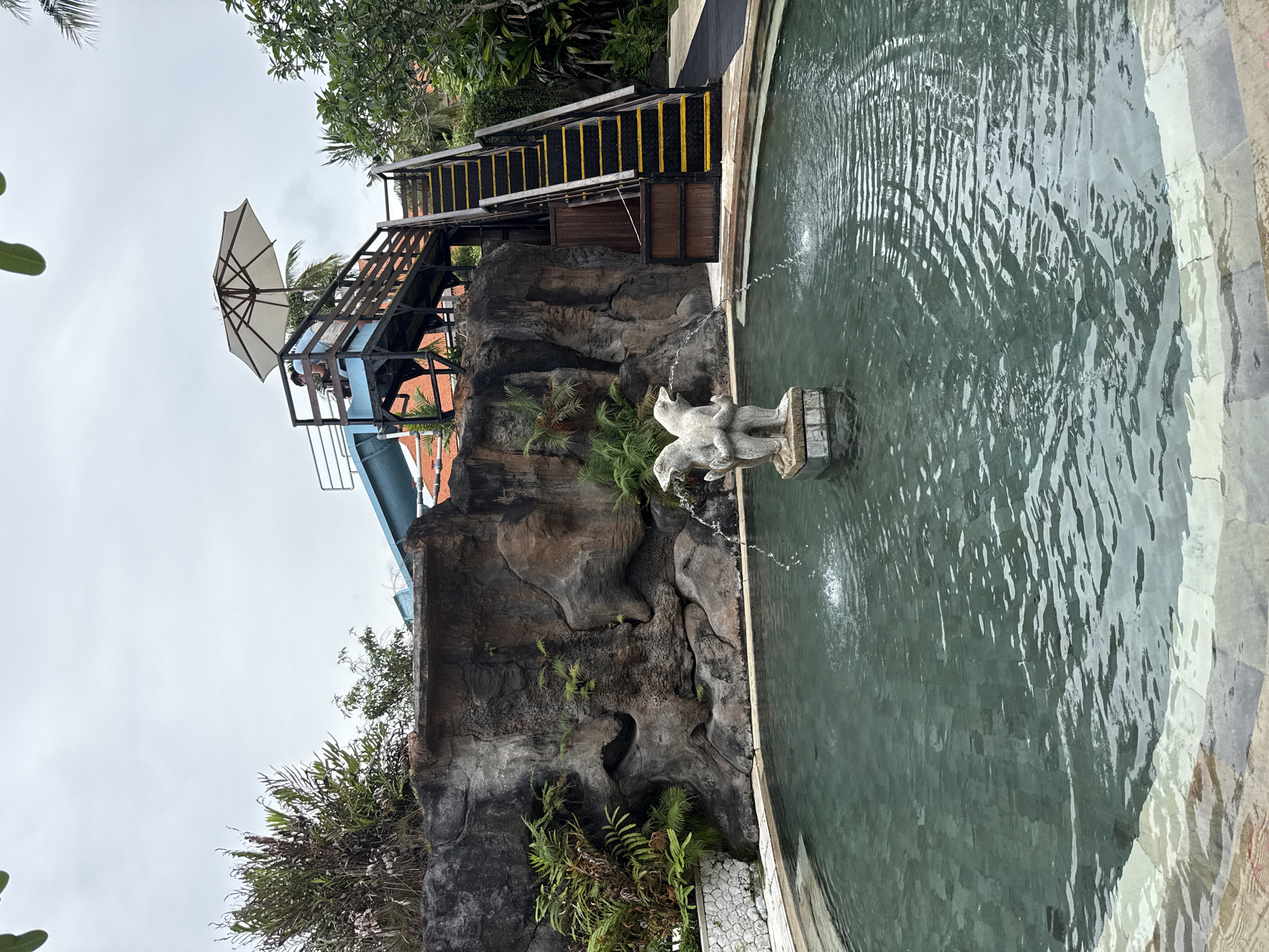 The image shows a water feature with a stone sculpture in a pool of green water, located in Nusa Dua, Indonesia. The sculpture depicts two animals. Water is being expelled from the sculpture into the pool. Behind the water feature is a rocky wall with vegetation growing on it. To the left is the bottom of a water slide. To the right is a staircase with yellow-and-black accents, which leads to a platform. An umbrella is visible on the platform. The sky is overcast.