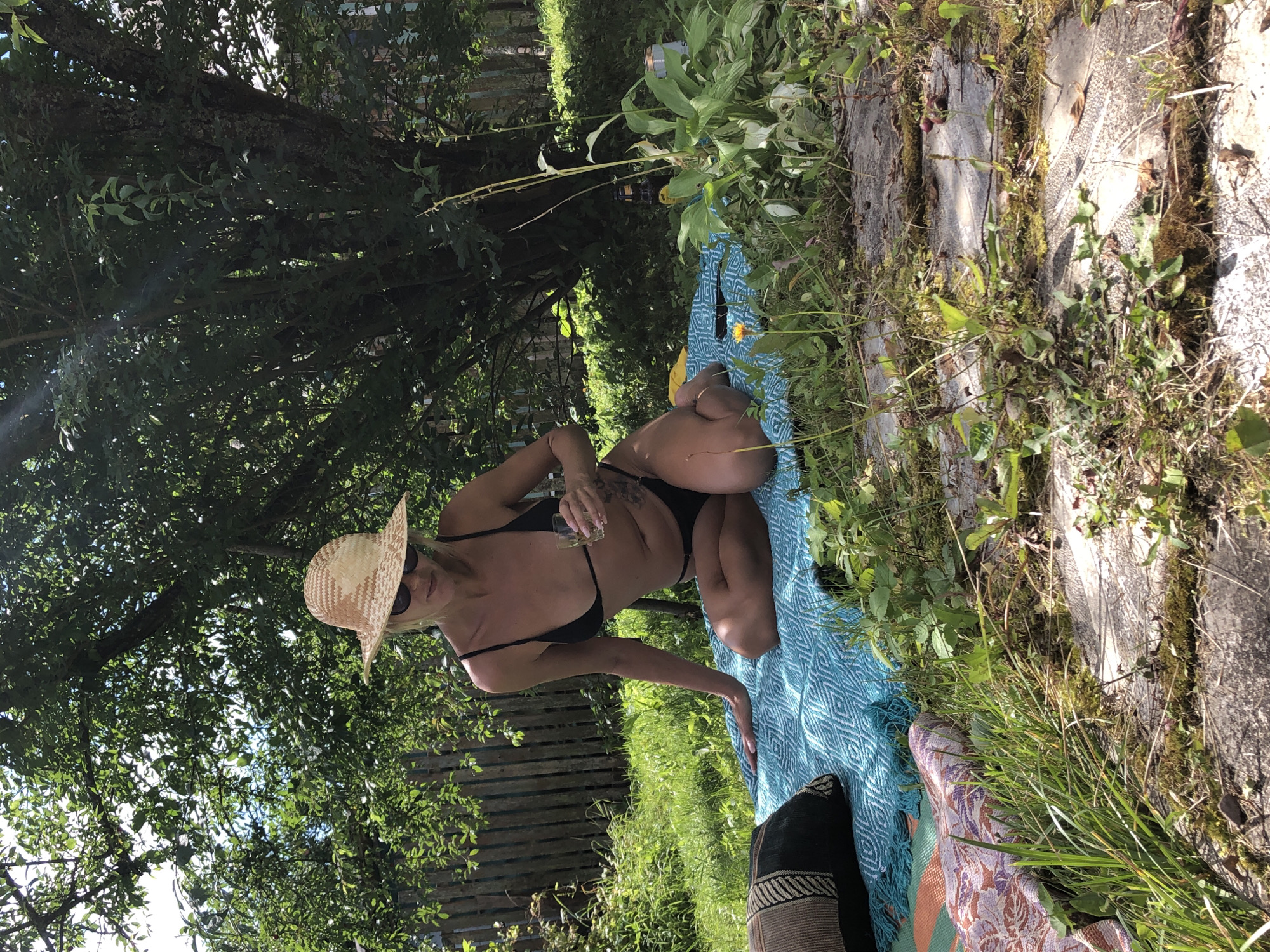 The image depicts a woman wearing a black bikini and a straw hat, sitting on a blue blanket in a garden. She is holding a glass of liquid and appears to be sunbathing. The background shows lush green trees and foliage, a wooden fence, and some grass. The overall setting suggests a relaxed outdoor environment, likely a private garden in Красная Пресня Снт, Russia, given the context.