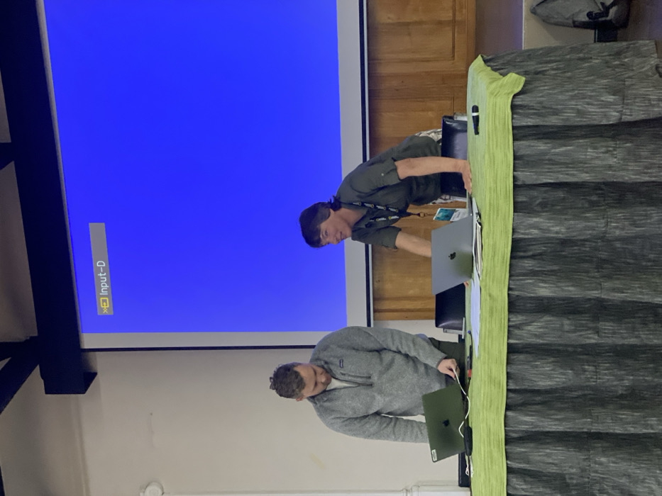 The image captures an indoor setting, likely a meeting or presentation room, in Riara Ridge, Kenya, featuring two men engaged with technology. A large projection screen dominates the left side of the frame, displaying a solid blue background with a white "Input-D" message and an 'X' symbol in the bottom left, indicating that a video input is not currently connected.
In the center, two men stand behind a table covered with a light green tablecloth and a darker green pleated skirt. The man on the left wears a grey quarter-zip sweater and is leaning over a dark-colored laptop, looking intently at its screen. The man on the right wears a dark green long-sleeved shirt and has a lanyard around his neck, bearing a yellow-bordered badge with faintly visible "NATIONAL GEOGRAPHIC" text. He is leaning over the table, seemingly interacting with a silver laptop or other items. Both laptops have the visible Apple logo.
A microphone and some documents are also present on the table. The background includes light-colored walls and wooden paneling. A black structural beam is visible in the upper left of the image. The scene suggests a professional environment, with the men focused on setting up or managing equipment for a presentation or event. The time of day is not discernible, but the space is well-lit.