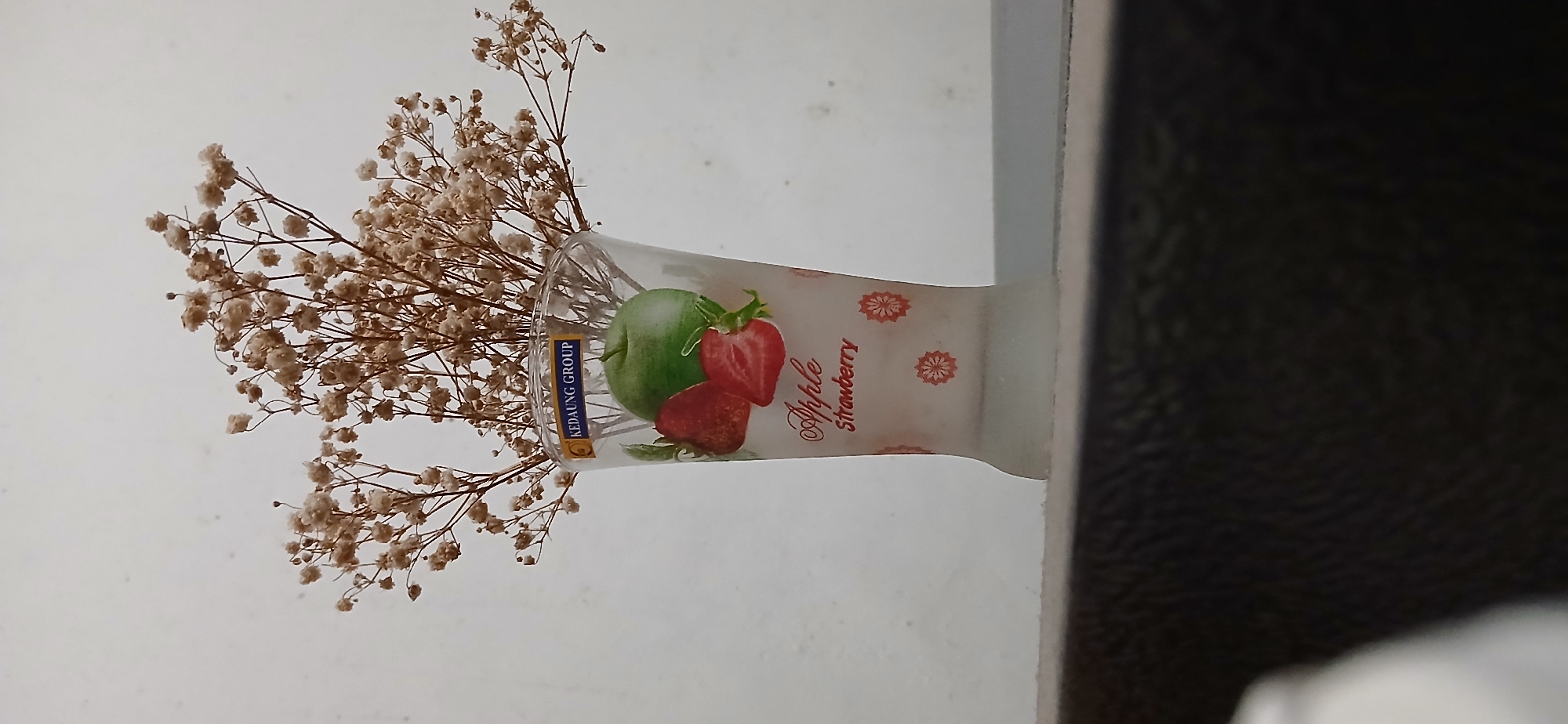 The image shows a clear glass vase filled with dried baby's breath flowers. The vase has a design featuring an apple, strawberries, and the words "Apple Strawberry" printed in red script. A blue and white label with the text "KEDAUNG GROUP" is affixed to the upper part of the vase. The vase is placed on a dark, textured surface, possibly a shelf or table, in front of a plain, light-colored wall. The lighting suggests an indoor setting. The image lacks any discernible indicators of a specific location within Samarinda, Indonesia. There are no people, actions, or interactions depicted.