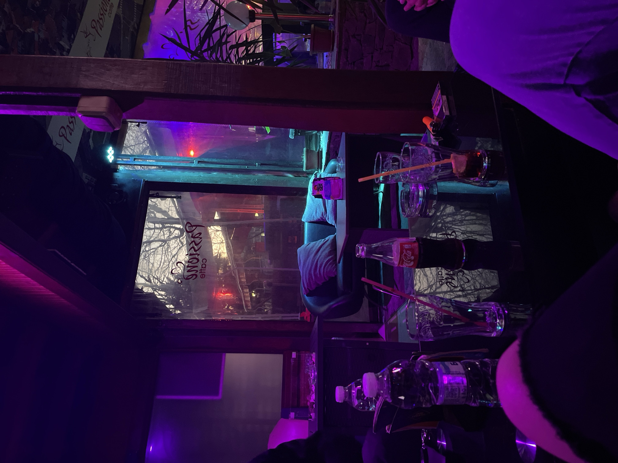 The image depicts an indoor scene in what appears to be a bar or café, evidenced by the presence of bottles of water, Coca-Cola, and glasses. The interior is illuminated with purple and blue lighting, creating a modern and stylized atmosphere. There is a window, possibly a storefront window, showing the exterior view. The reflection on the glass suggests the business is "Passione Caffe", which could possibly indicate a location of Zlatograd, Bulgaria. Two persons' clothing can be seen along the right edge of the image. The environment is dimly lit.