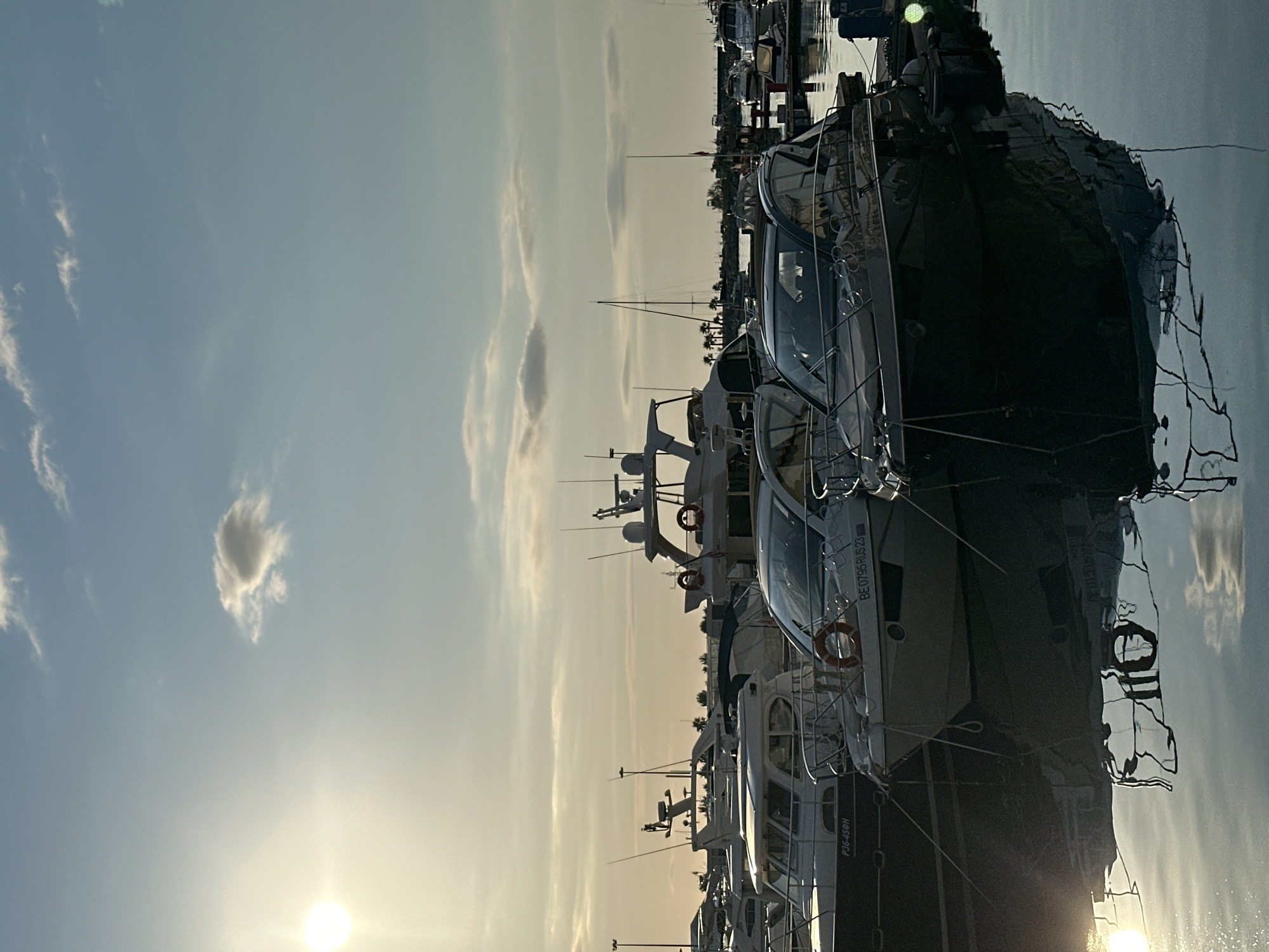 This is a photograph of a marina filled with several yachts, viewed from a low angle. The sun is setting or rising, casting a warm glow across the sky, which is filled with wispy clouds. The water in the marina is calm and reflects the yachts and the sky, creating a mirror-like effect. The overall mood is peaceful and serene.
The primary subjects are the yachts themselves, which are modern and well-maintained. They are docked side-by-side, suggesting a busy but orderly marina. Some of the yachts have life preservers visible, which are orange and circular. There are no people clearly visible in the photograph, but their presence is implied by the docked boats.
The setting is a marina, likely in Sochi, Russia, given the context. The time of day is either sunrise or sunset, indicated by the low angle of the sun and the warm, diffused light. The weather appears to be calm and clear, with only a few clouds in the sky. The water is still, suggesting a lack of wind.
Notable details include the reflections of the yachts and sky in the water, which are distorted but still recognizable. The sky has a gradient of colors, from a pale blue at the top to a warmer yellow and orange near the horizon. There is a bright, circular light source, presumably the sun, low in the sky.
Visible text includes "BE 0795 RUS 23" and "P36-450H" on the sides of the yachts. This text likely refers to registration numbers or identifiers for the vessels. The photograph captures a moment of quiet beauty in a popular maritime location.