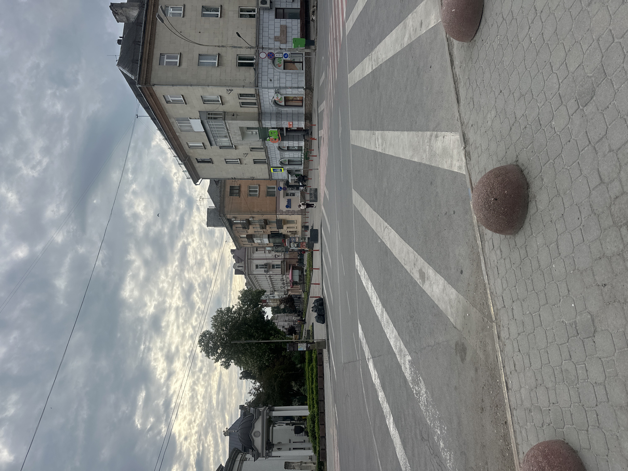 The image shows a street in Ternopil, Ukraine. In the foreground, there's a pedestrian crossing with broad white stripes painted on the road. Adjacent to the crossing are rounded, reddish-brown bollards placed on a sidewalk paved with gray bricks. Buildings line the left side of the street, varying in height and architectural style, appearing to be multi-story structures. Some have balconies and visible windows. The sky above is filled with layered clouds, creating a diffused light. Further down the street, trees and other buildings are visible, along with hints of pedestrian and vehicle activity.