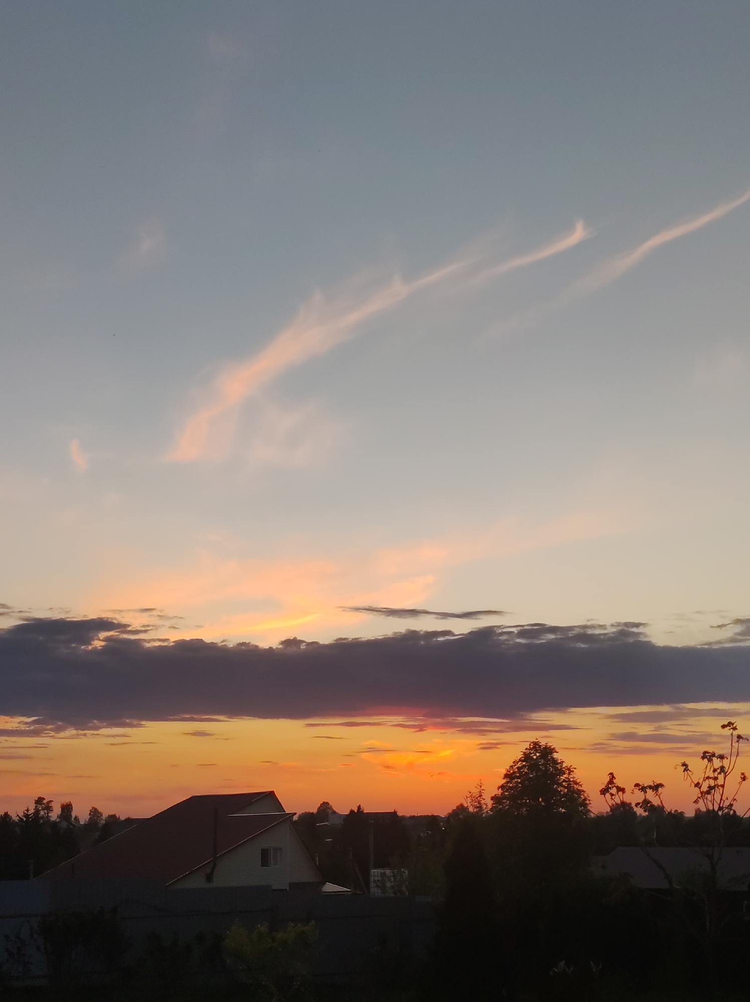 The image captures a sunset over a suburban area, possibly near Фоки, Russia. In the foreground are the silhouettes of houses and trees, partly obscured by a fence and shrubbery. Above them, the sky is ablaze with orange and red hues, transitioning into a band of dark, layered clouds. Higher up, the sky fades into lighter shades of pink and blue, with long, thin wisps of clouds stretching across the upper part of the image.