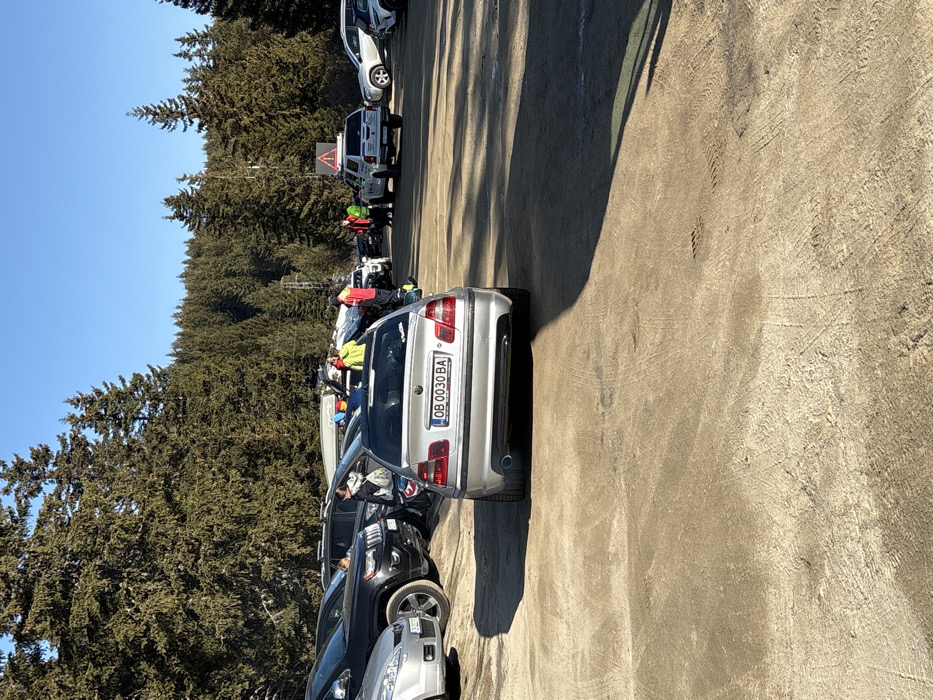 The image displays an unpaved outdoor parking area situated within a dense evergreen forest in a mountainous region of Bistritsa, Bulgaria, under a clear blue sky. In the foreground, a silver BMW E46 sedan with Bulgarian license plate OB 0030 BA is parked facing away from the viewer. The ground is light brown, compacted dirt or gravel, exhibiting various tire tracks and long, distinct shadows.
In the midground, multiple vehicles are parked. These include a black Jeep Grand Cherokee and a silver hatchback visible to the left of the BMW. Further back and to the right, a white pickup truck, possibly a Toyota Hilux, is parked. This truck features a roof-mounted triangular warning sign with an exclamation mark, and a similar warning sign is positioned on a pole immediately behind it. A group of approximately five to seven individuals, clad in winter clothing including bright yellow and red jackets, are congregated near the parked vehicles, appearing to engage in conversation or preparation. Some vehicles have items secured to their roofs, such as a white watercraft resembling a kayak or canoe.
The background is dominated by a thick forest of tall evergreen trees covering a mountain slope under the bright blue sky. A power line structure is partially discernible within the trees in the mid-to-background.
