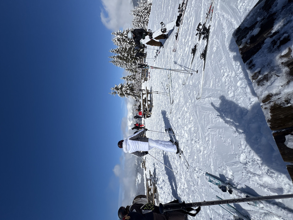 The image depicts a high-altitude plateau or summit within the ski resort of La Clusaz, France, on a clear, sunny winter day. The ground is entirely covered in granular snow, showing tracks from skis and footsteps. The sky is a deep, cloudless blue overhead, with some white, scattered clouds visible along the distant horizon.
In the foreground, leaning against an unseen object on the left, is a pair of grey ski poles with blue accents. Below them, two pairs of skis lie flat on the snow. In the lower right, the snow-dusted edge of a rough wooden structure, possibly a table or bench, is visible.
Multiple individuals, dressed in winter ski attire, are present. In the mid-ground, slightly left of center, a person wearing a white ski jacket with grey shoulders, white ski pants, and a dark helmet stands on skis, holding ski poles, facing away from the camera towards the distant landscape. To their right, a person in a dark jacket and helmet holds a white snowboard vertically. Several other pairs of skis and poles are resting on the snow between these individuals.
Further into the mid-ground and background, multiple wooden picnic tables with benches are distributed across the snow, some partially covered. Several individuals are visible around these tables; some appear to be seated or standing, dressed in various colored ski jackets, including red and dark tones. Behind the picnic tables, a dense cluster of snow-laden evergreen trees forms a natural windbreak or landscape feature. In the far background, snow-capped mountain peaks and a valley are discernible under the clear sky. Long shadows of people and objects are cast upon the snow, indicating bright sunlight.