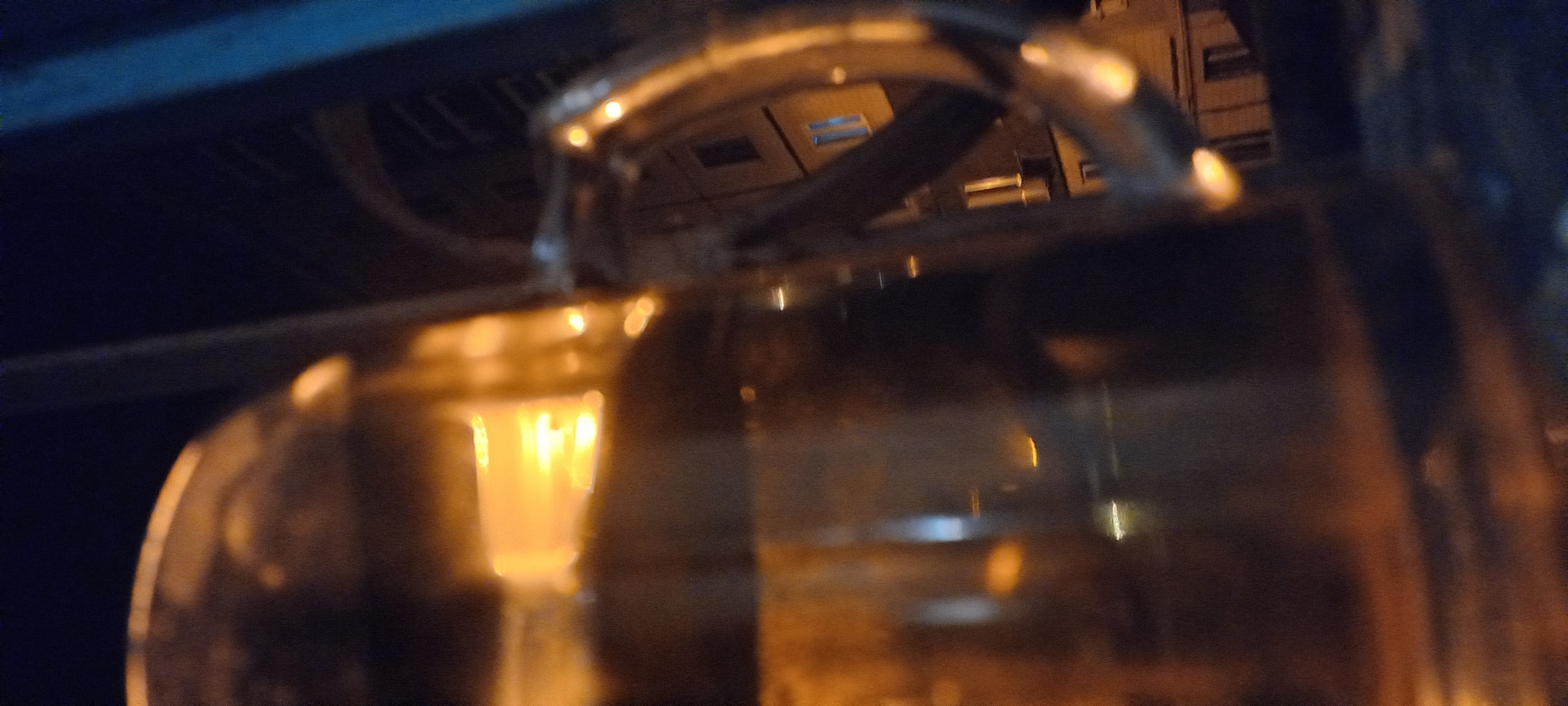 The image depicts a blurred nighttime scene, likely captured in Kryvyi Rih, Ukraine. The foreground is dominated by a large, translucent object, possibly a glass container or a window, with warm, orange light reflecting off its surface. Beyond this object, a dark, out-of-focus background suggests an urban environment. There are hints of buildings with lit windows, indicated by rectangular shapes with soft yellow glows. A vertical element, potentially a pole or railing, is visible on the right side of the frame, with a blueish tint. The overall impression is one of ambient city lighting at night, seen through a reflective or translucent medium. No people or specific actions are discernible due to the blurriness and lighting conditions.