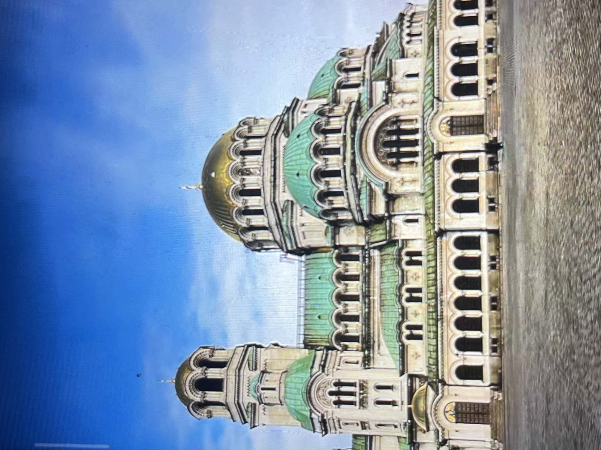 The image shows the St. Alexander Nevsky Cathedral in Sofia, Bulgaria, rotated 90 degrees counter-clockwise. The cathedral, a large Eastern Orthodox basilica, is visible against a blue sky. Its architectural details, including the golden dome, green copper domes, arched windows, and ornate facade, are clearly depicted. The ground or plaza surrounding the cathedral is visible on the right. The location is NOT Tinaquillo, Venezuela.