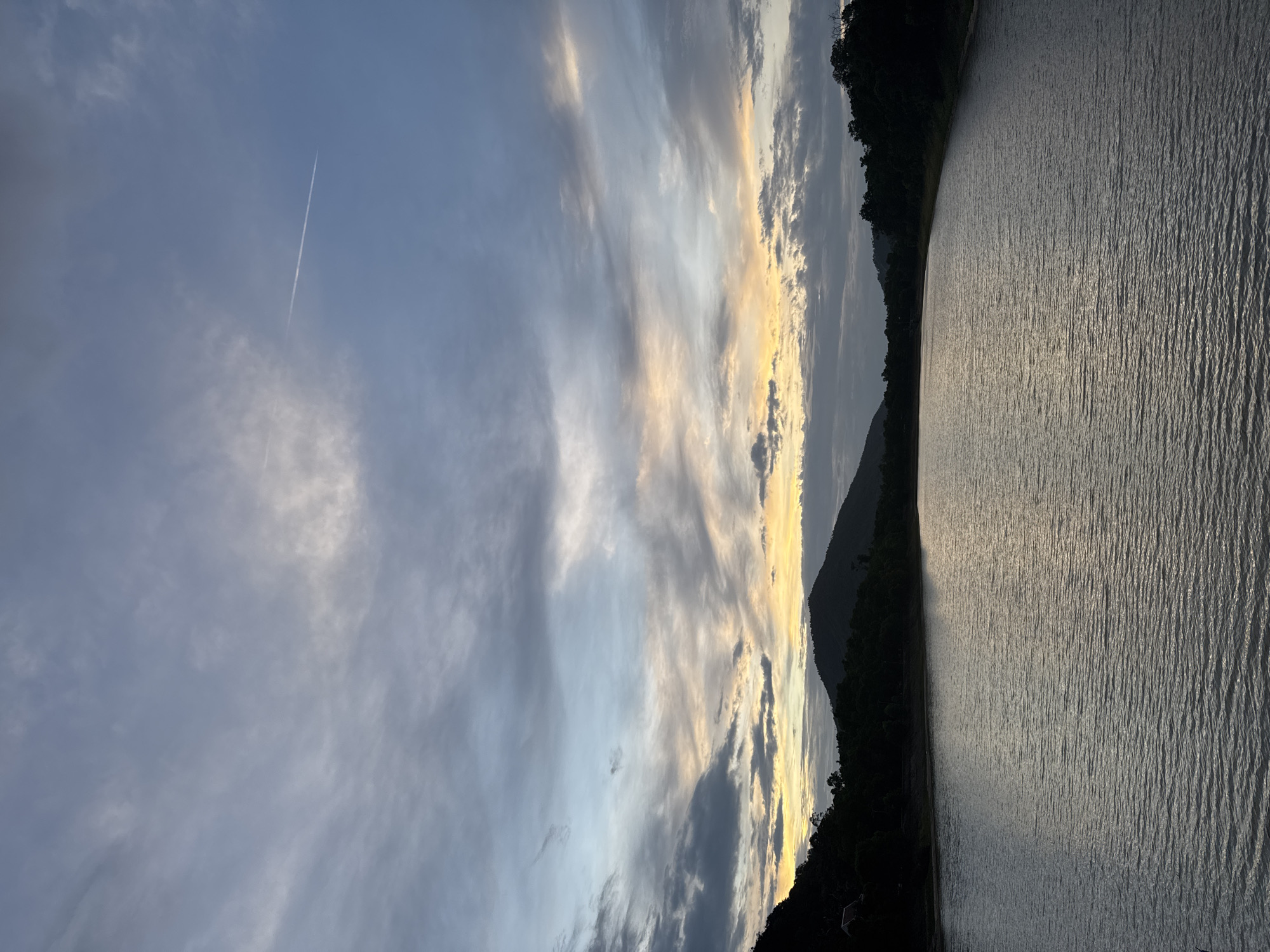 An outdoor, vertical, medium shot captures a serene body of water with a backdrop of silhouetted hills and a dramatic cloudy sky. The water's surface is rippled, reflecting the muted colors of the sky, creating a textured, silvery sheen. The far shore is lined with dark, dense foliage, with a prominent, rounded hill in the center, and two smaller, rounded hills to its left and right. A single, faint contrail from an airplane stretches diagonally across the upper right quadrant of the sky. The sky itself is a complex canvas of grays, whites, and soft yellows, indicative of either sunrise or sunset. The overall impression is one of quiet solitude and natural beauty. This scene is likely located near the Chaweng Lake in the Ko Samui District of Thailand, known for its picturesque landscapes and tranquil reservoirs that often feature similar mountainous terrains and vibrant skies. No people or specific actions are depicted in the image.