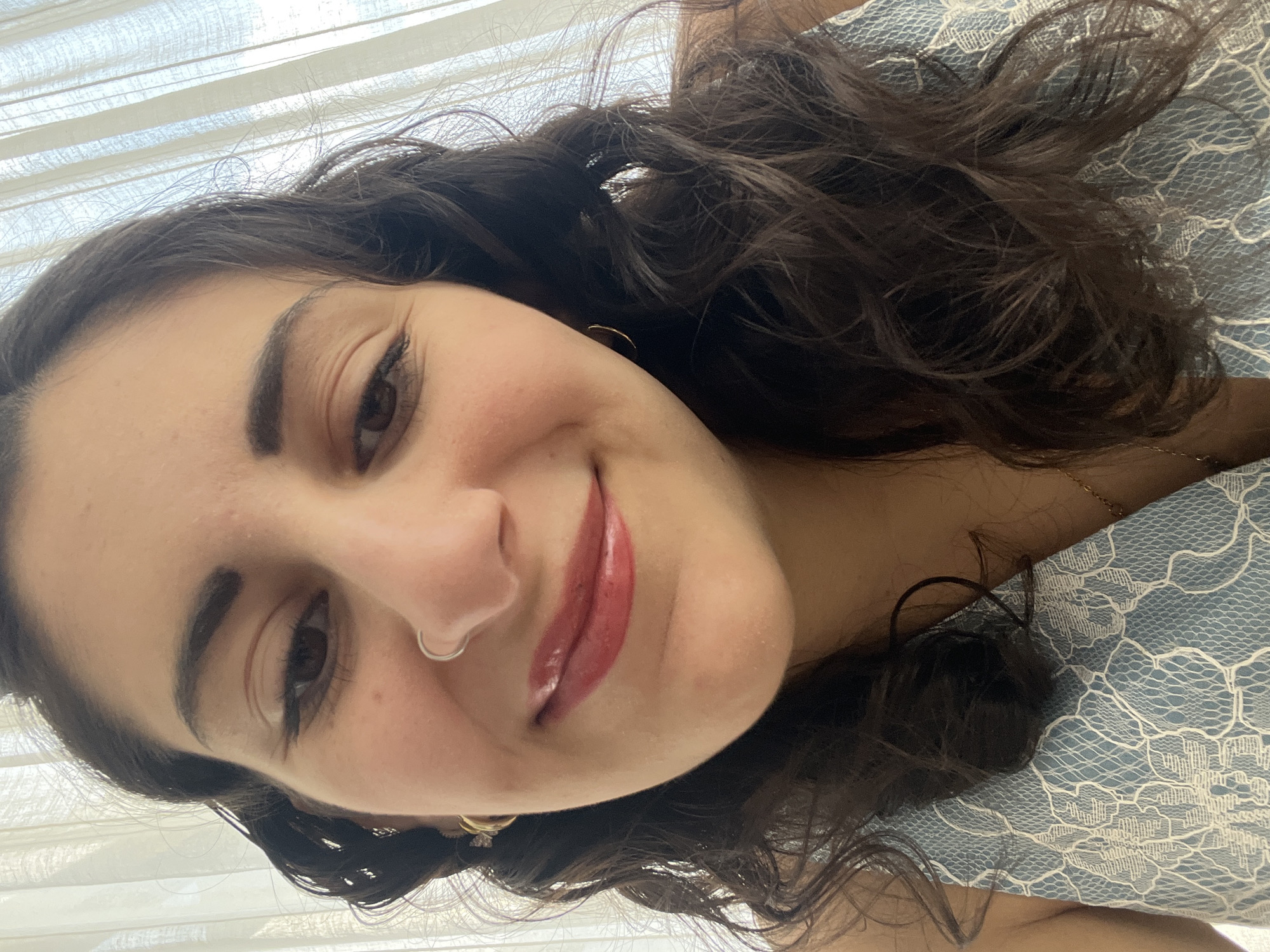 A close-up, selfie-style shot shows a woman with dark, curly hair, fair skin, and dark eyes. She is smiling and looking towards the camera. She has a nose ring and earrings. The background includes white curtains and a blue and white patterned surface, possibly a bedspread or piece of clothing. There is no information in the image to indicate this was taken in Mexico City, Mexico.